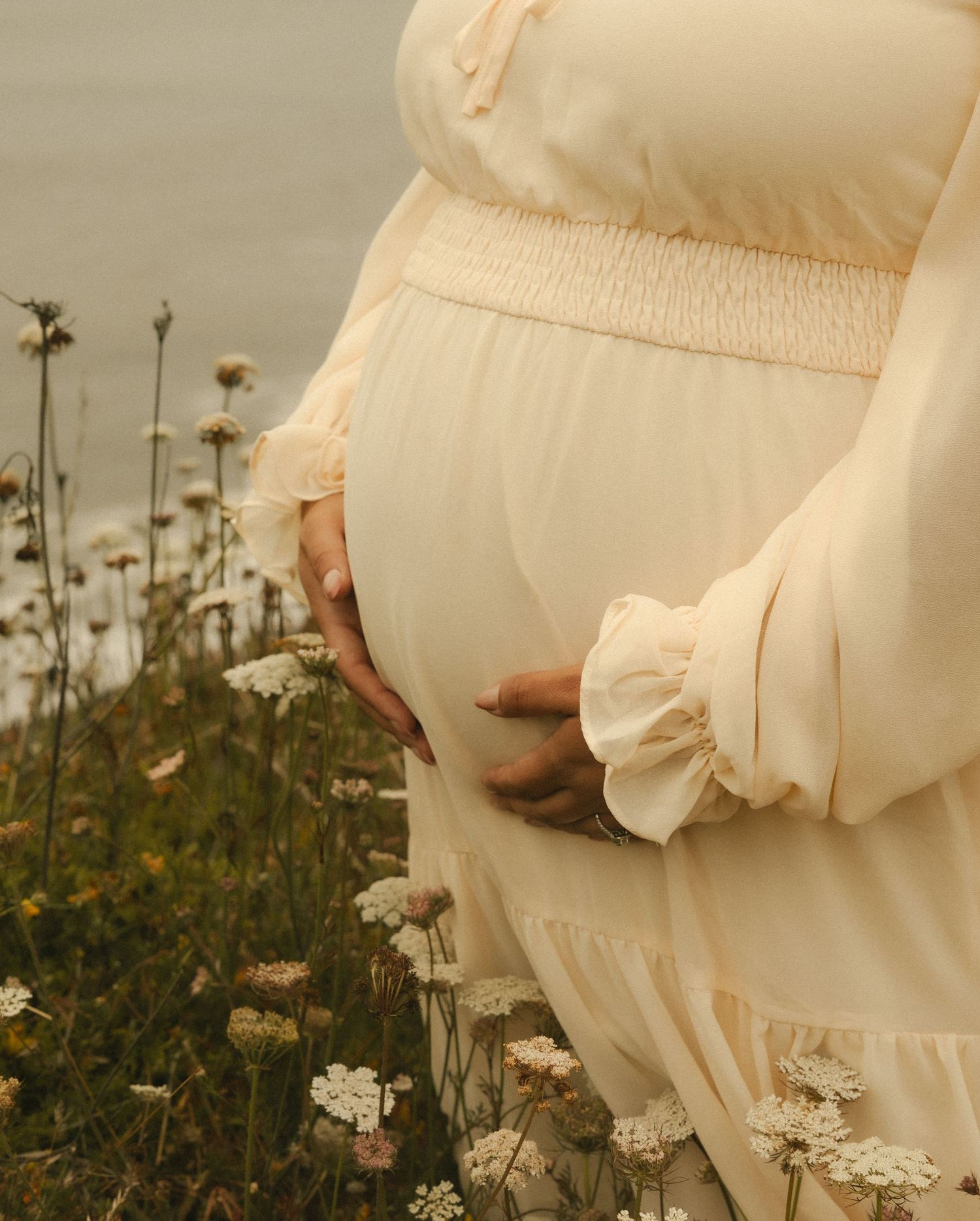 Pregnant woman in cream dress cradling baby bump among wildflowers in a serene outdoor setting.