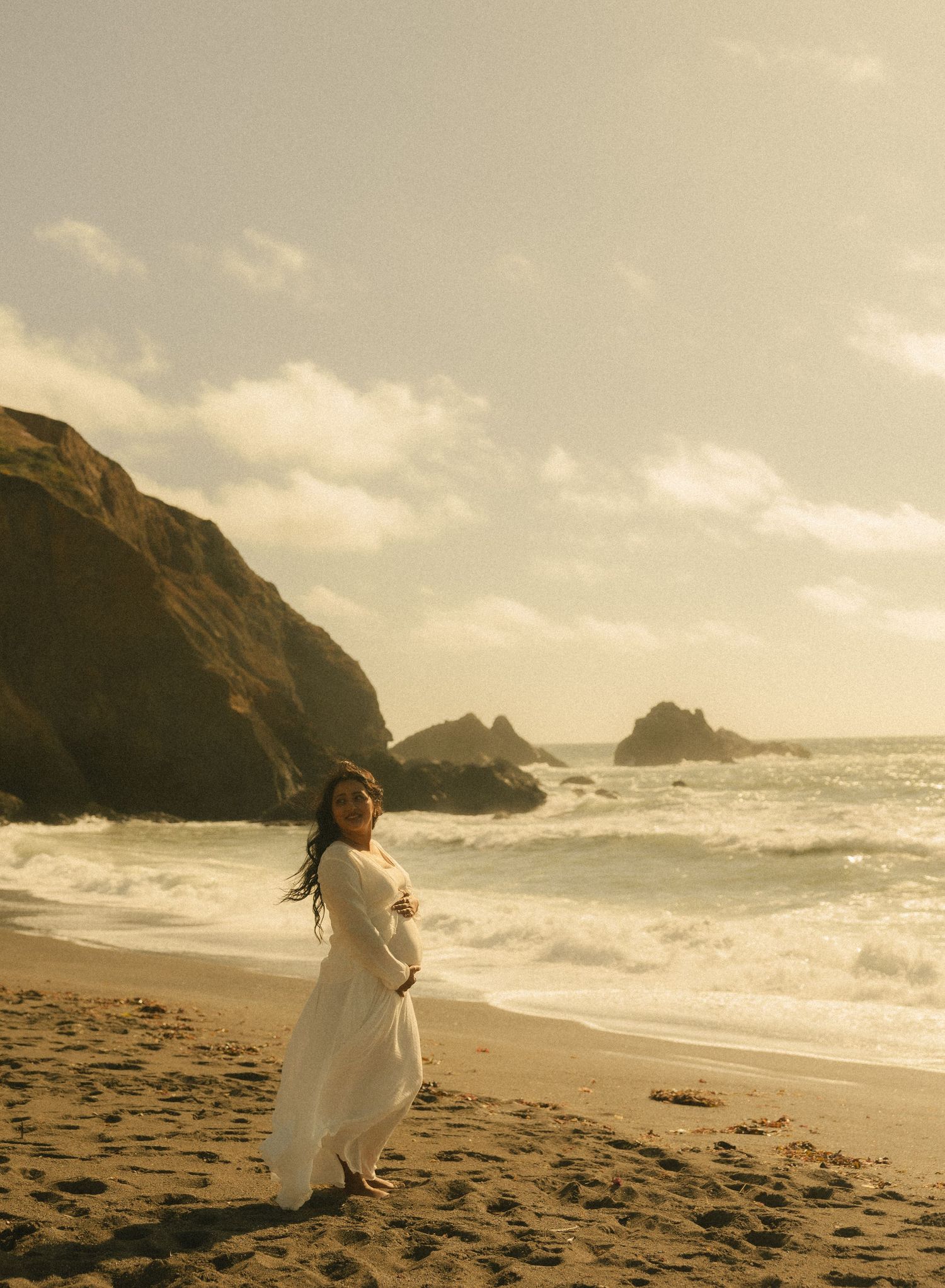 Pregnant woman in white dress on sandy beach with rocky cliffs and ocean waves in golden light.