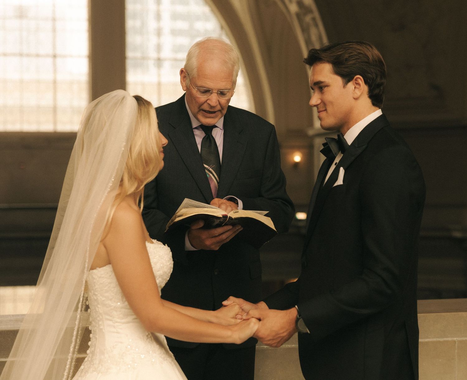 A bride and groom exchange vows before an officiant holding a book during a formal wedding ceremony.