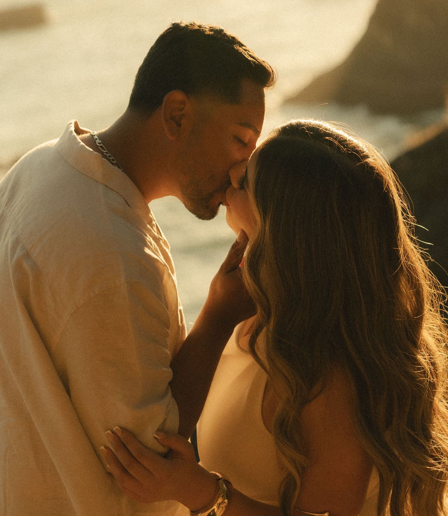 Romantic couple sharing an intimate moment at golden hour on a rocky beach, dressed in neutral tones.