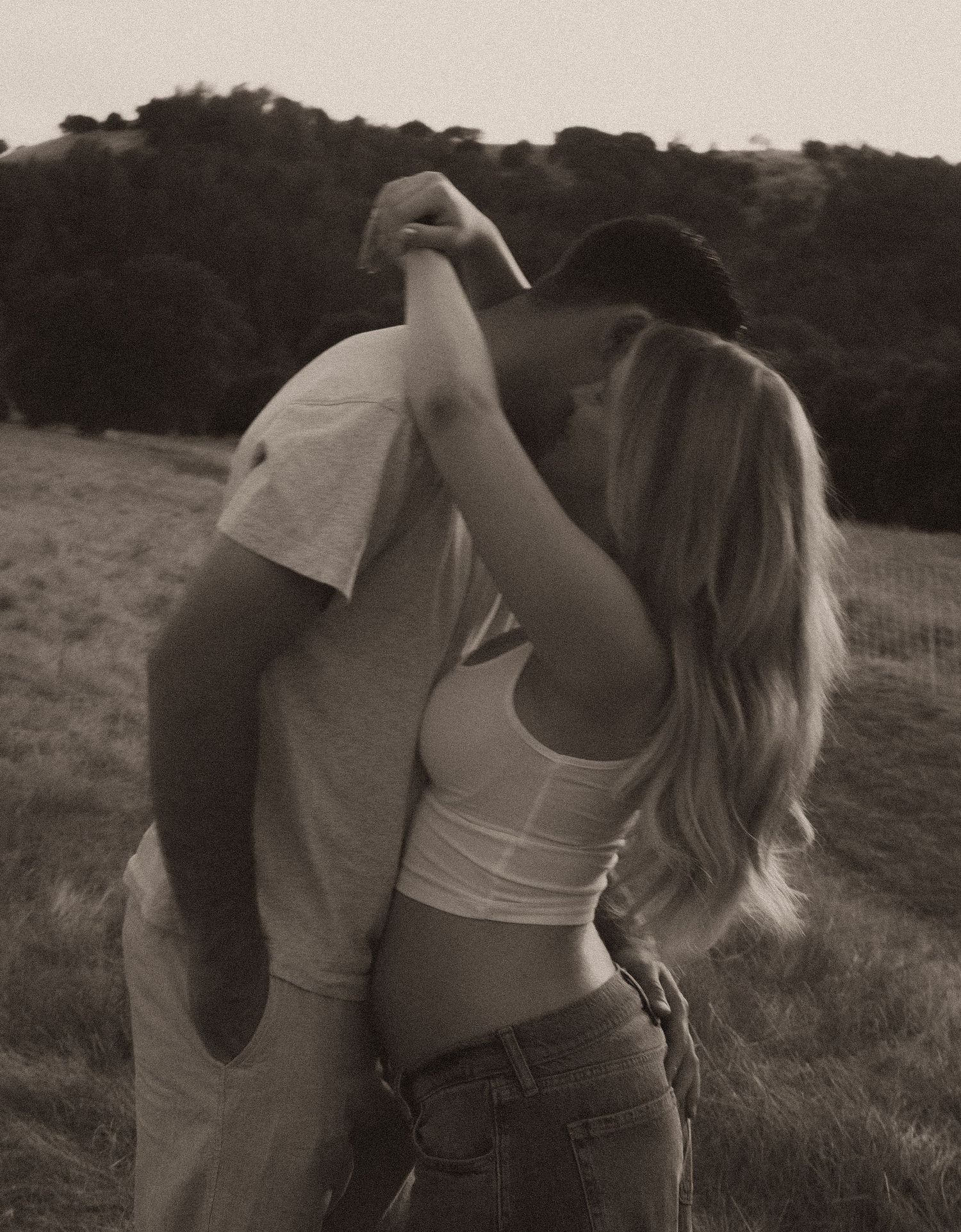 Romantic couple kissing and dancing in a field, black and white photo with rolling hills in background.