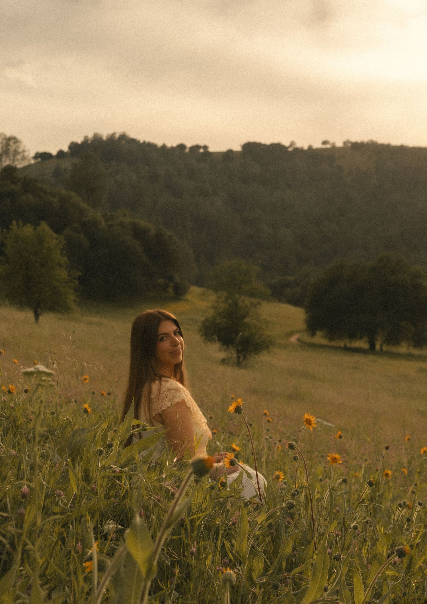 Young woman in white dress sitting among wildflowers in a golden meadow with rolling hills at sunset.