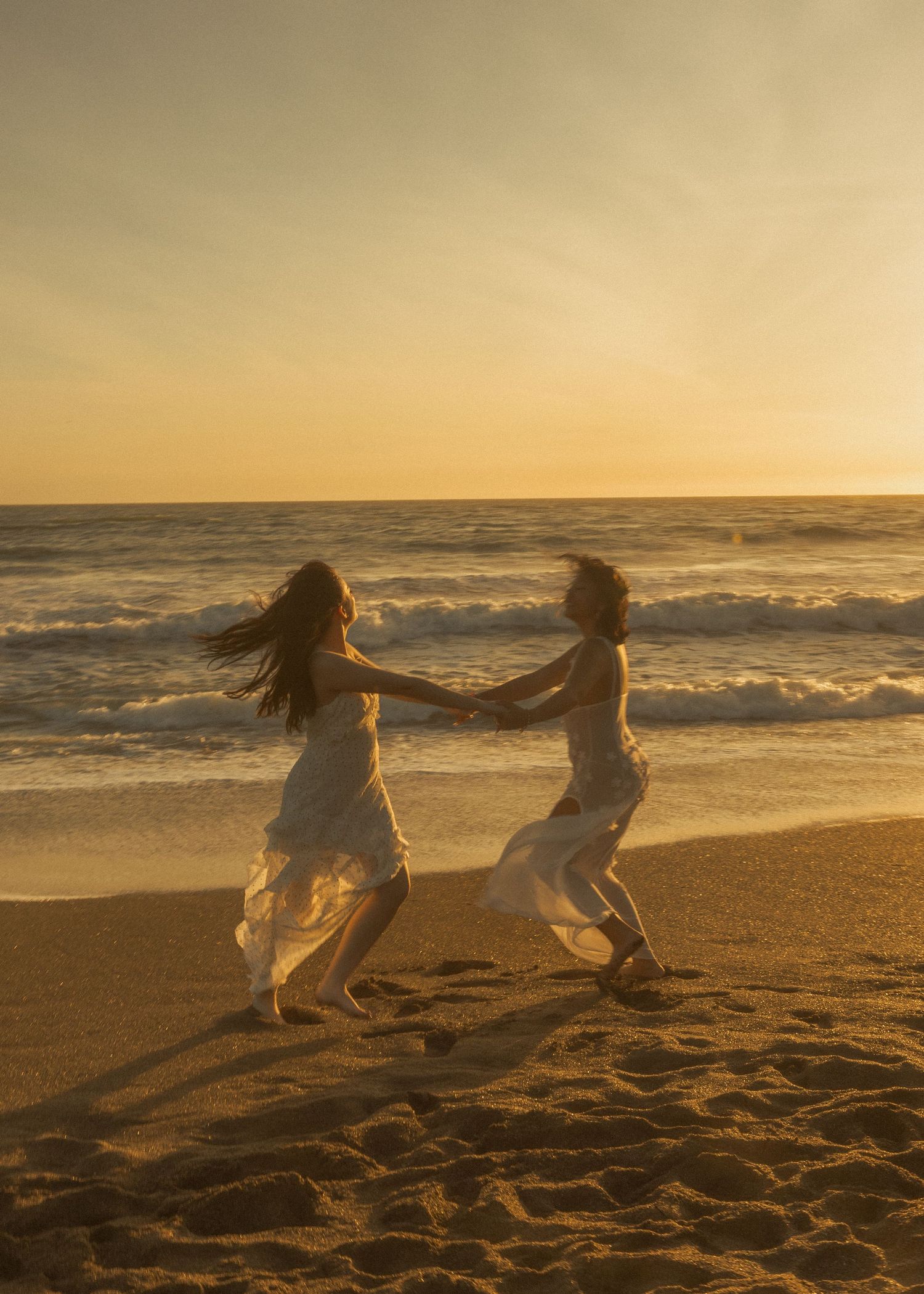 Silhouettes of people in flowing dresses twirl on a golden beach at sunset.