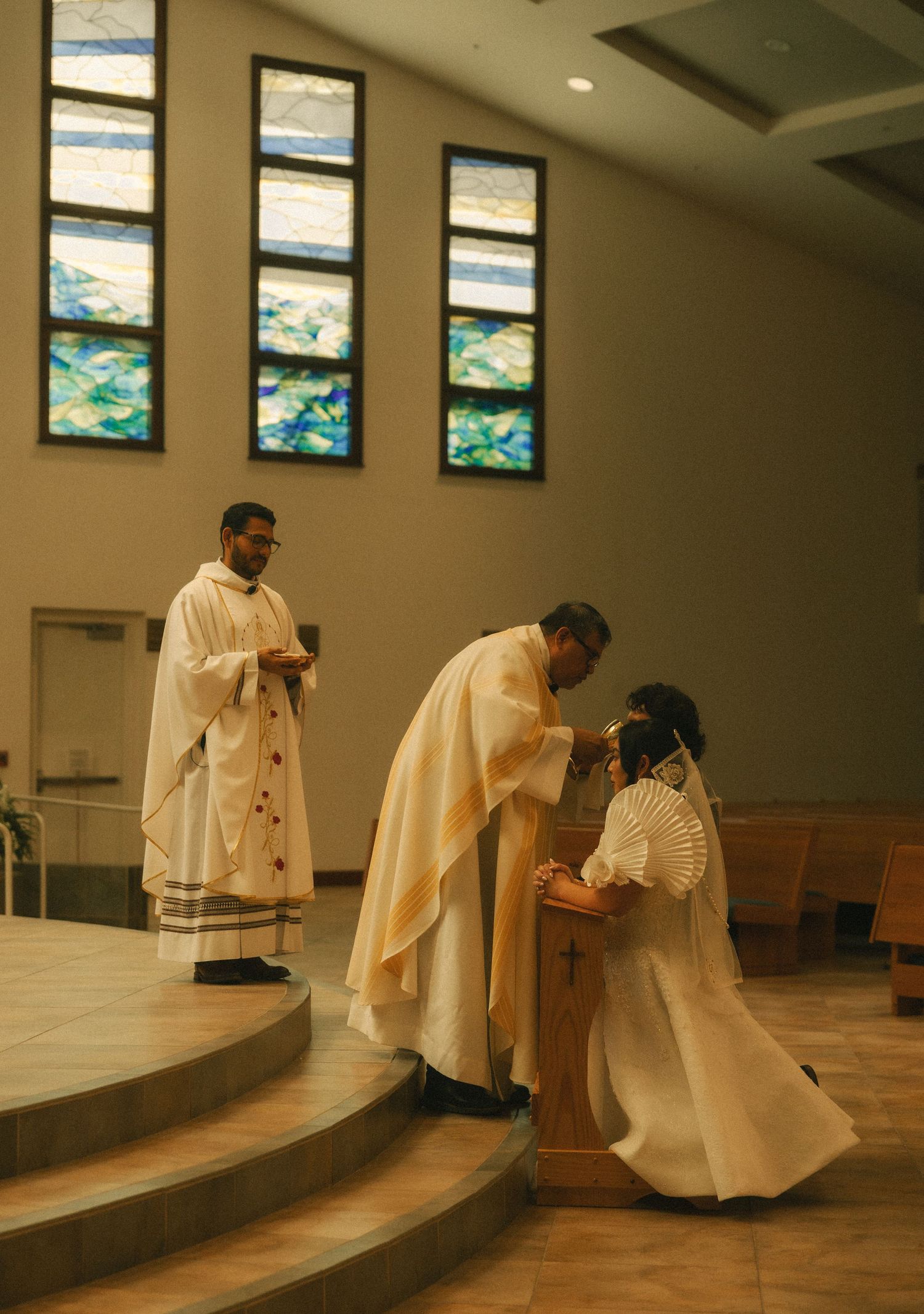 Priest blessing a kneeling person during a Catholic church ceremony with stained glass windows in background.
