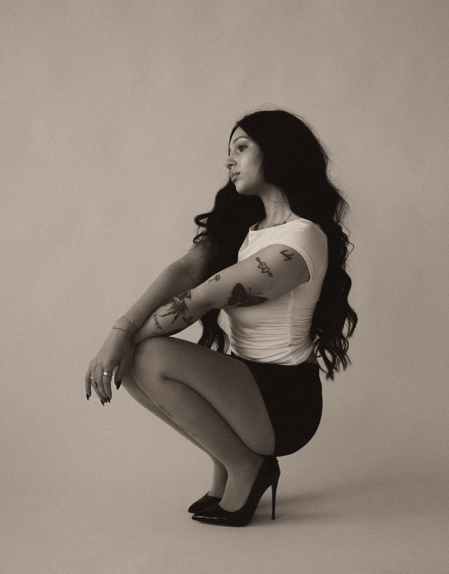 Black and white photo of a woman with long dark hair, tattoos, white top, squatting in heels.