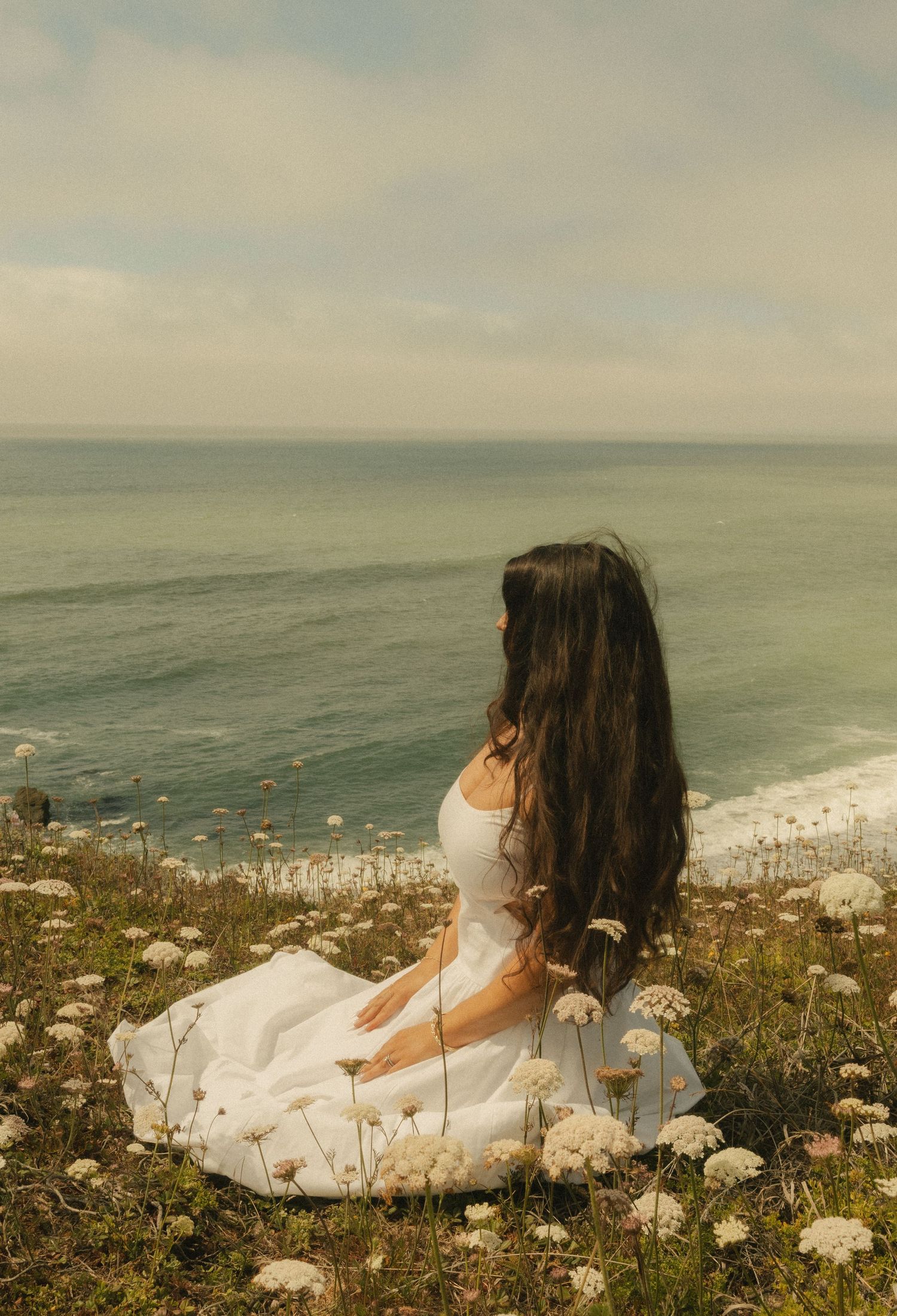 Woman in white dress sitting among wildflowers on coastal cliff, gazing at ocean waves under cloudy sky.