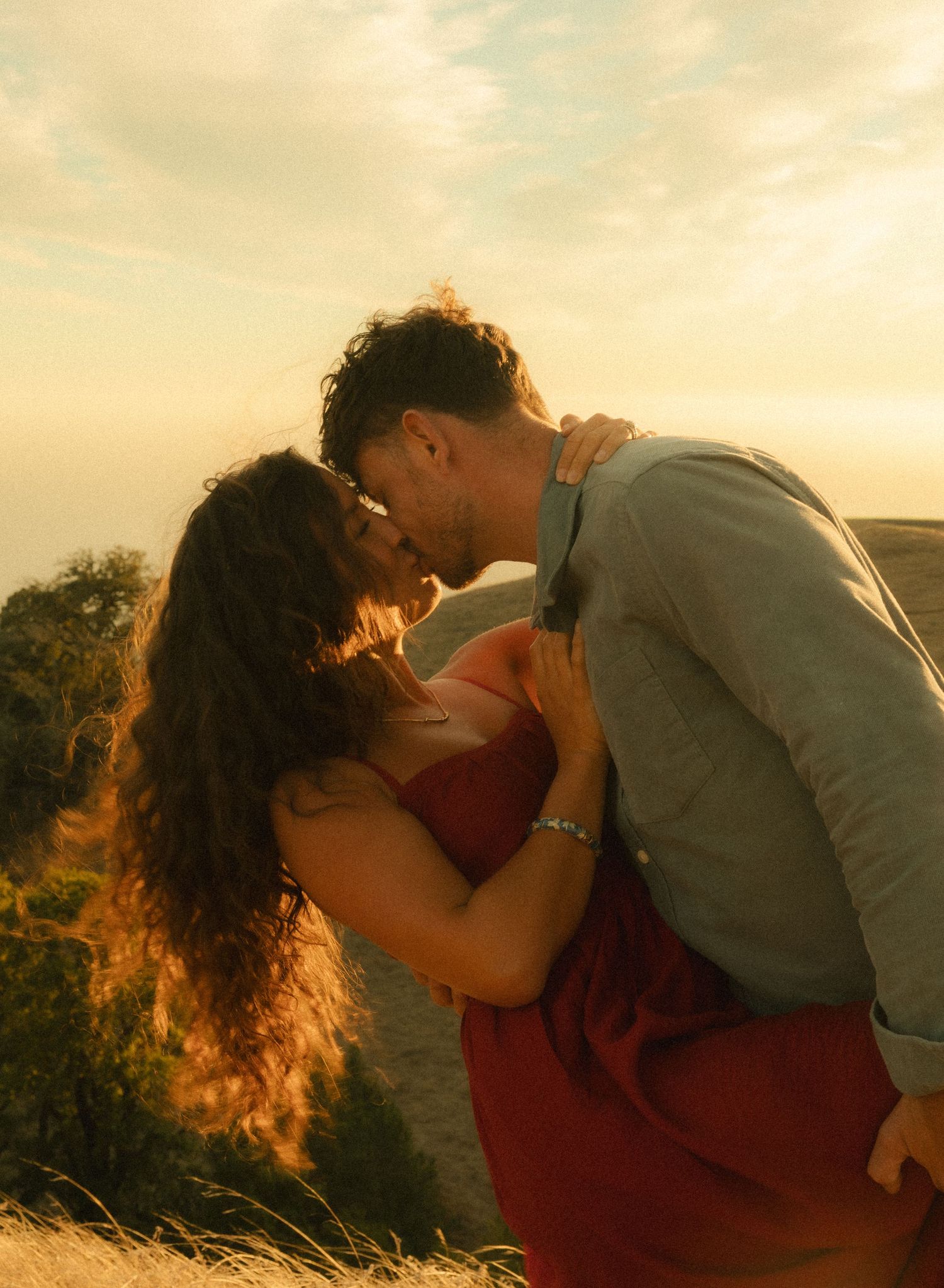 Couple sharing a romantic kiss at golden hour sunset on a hilltop, woman in red dress with curly hair.