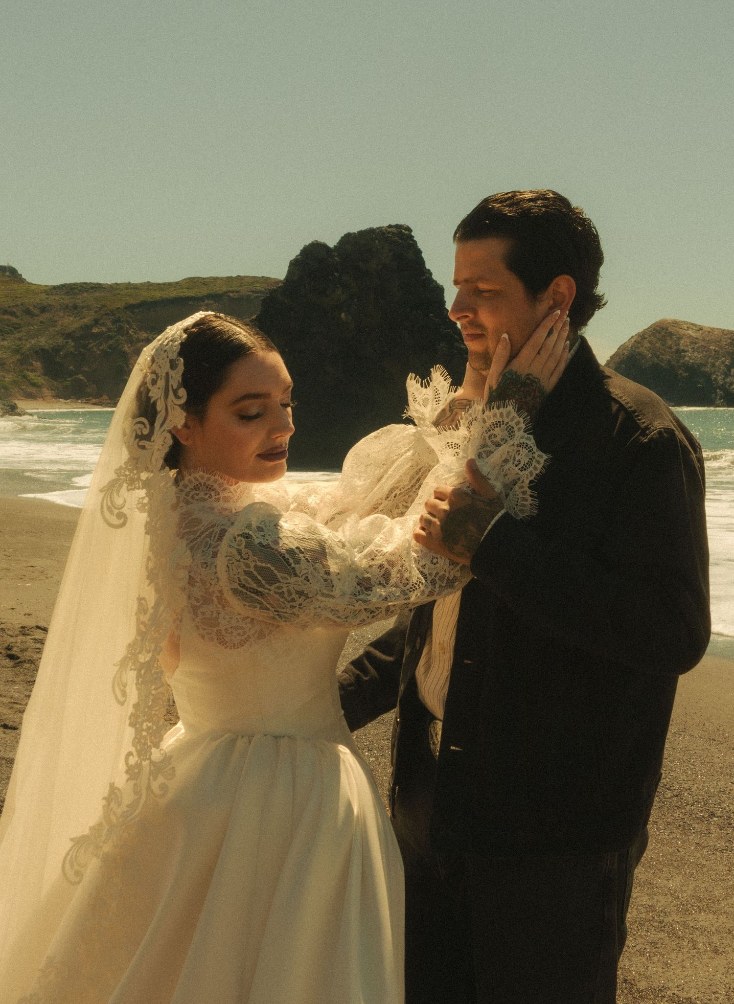 Bride in lace wedding gown and veil with groom in black suit on rocky beach shoreline.