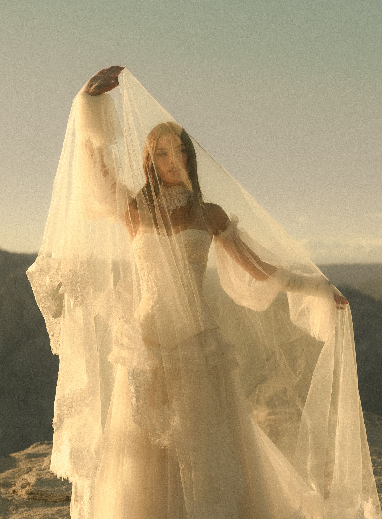 Bride in lace wedding gown lifting flowing veil against scenic mountain backdrop outdoors.