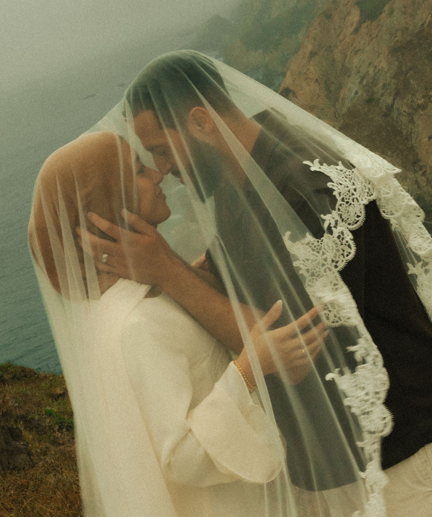Bride and groom sharing a romantic kiss under a lace-trimmed wedding veil with ocean cliffs in the background.