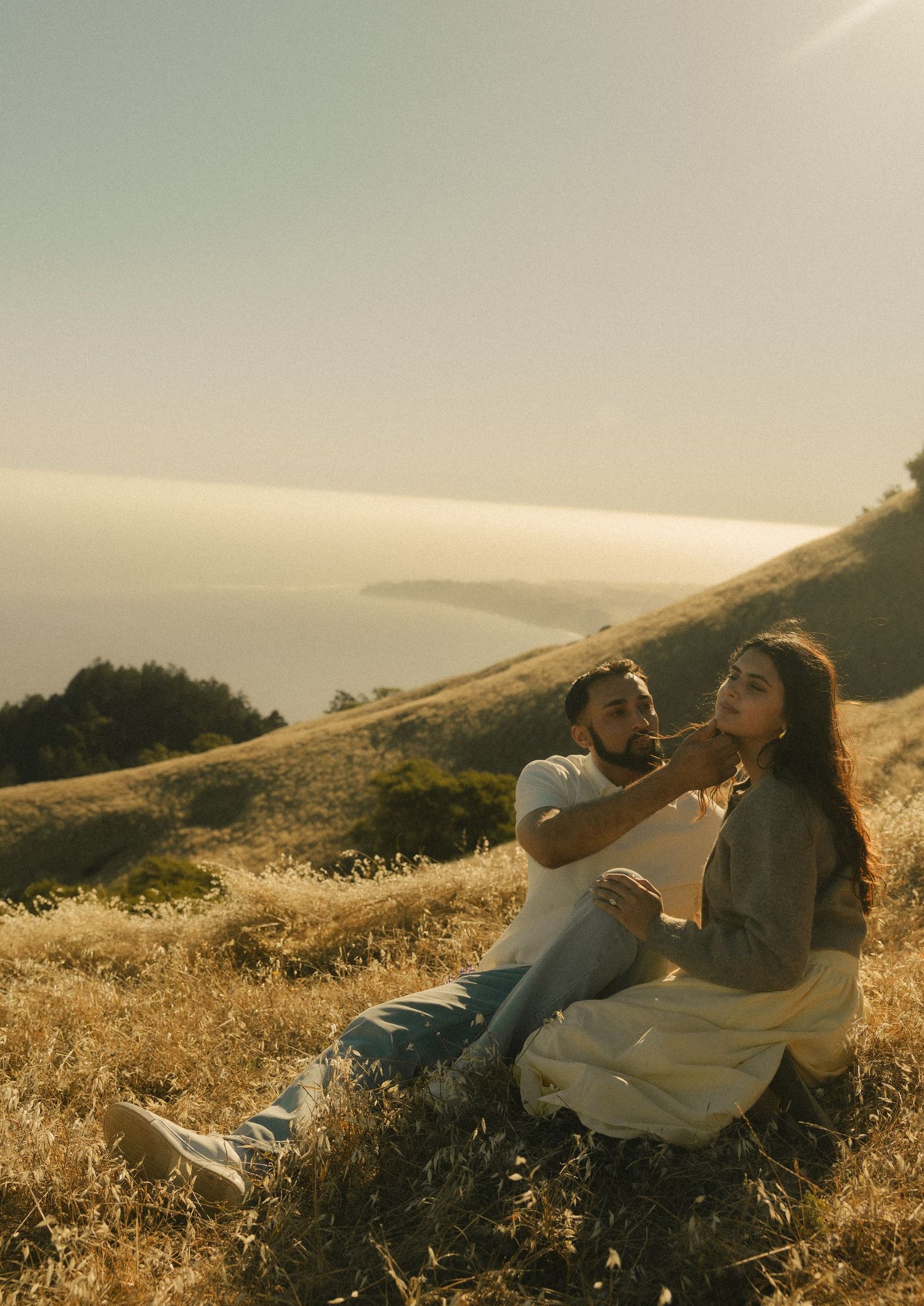 Couple sitting on golden hillside overlooking coastal ocean view during golden hour sunset.