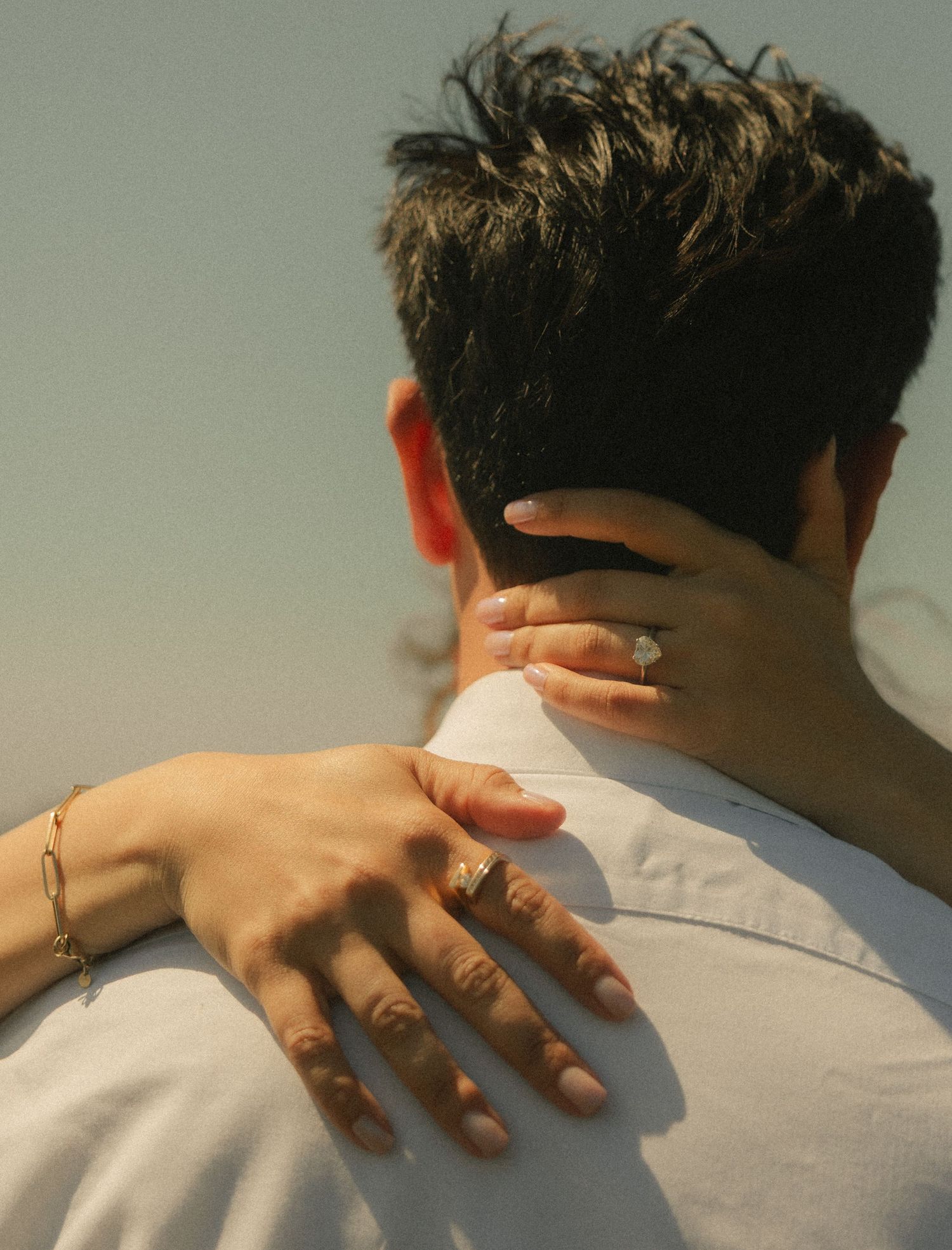 A woman's hands with an engagement ring embrace a man from behind against a soft sky backdrop.