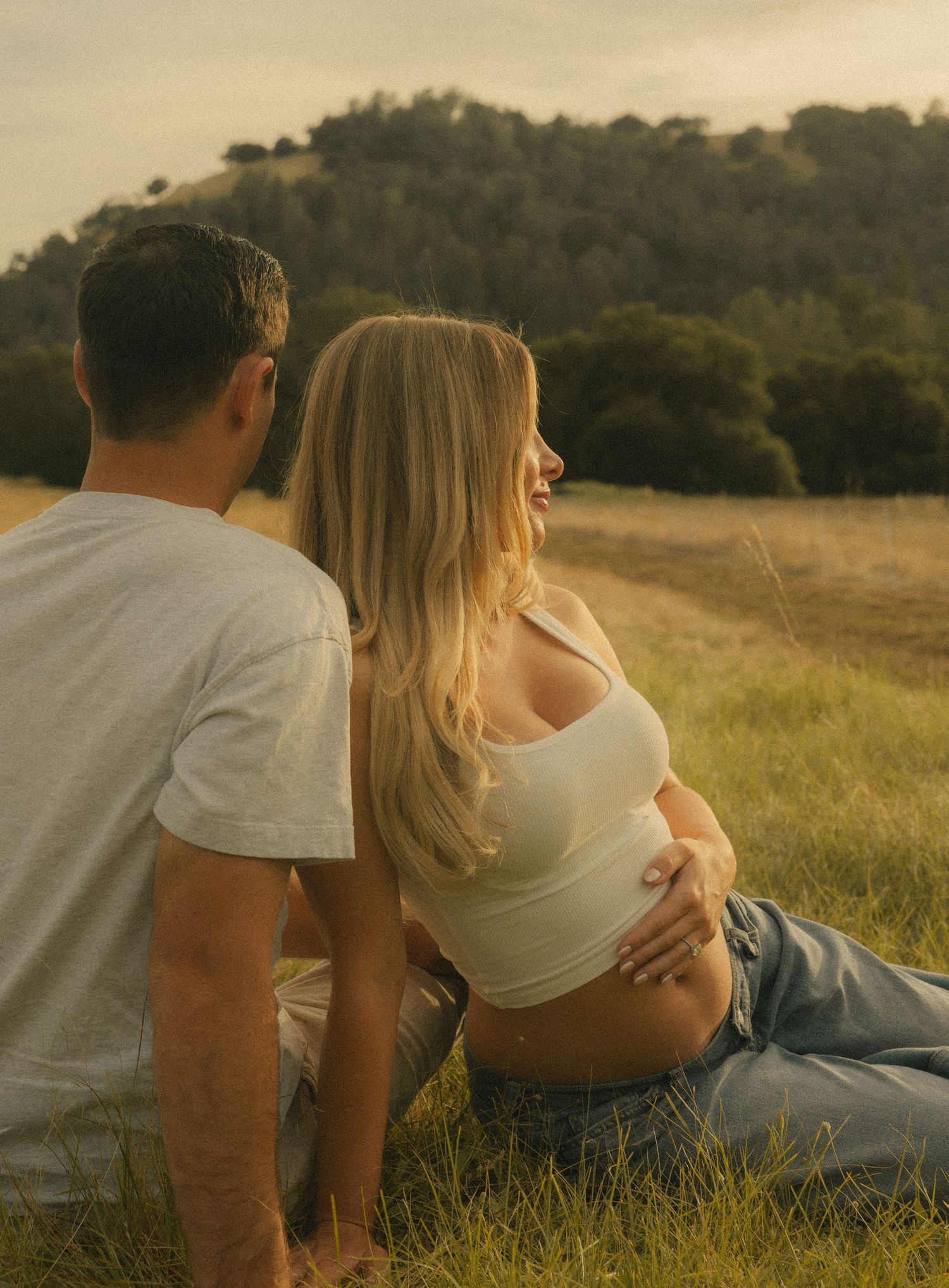 Pregnant woman in white crop top and jeans sits with partner in golden grass field at sunset.