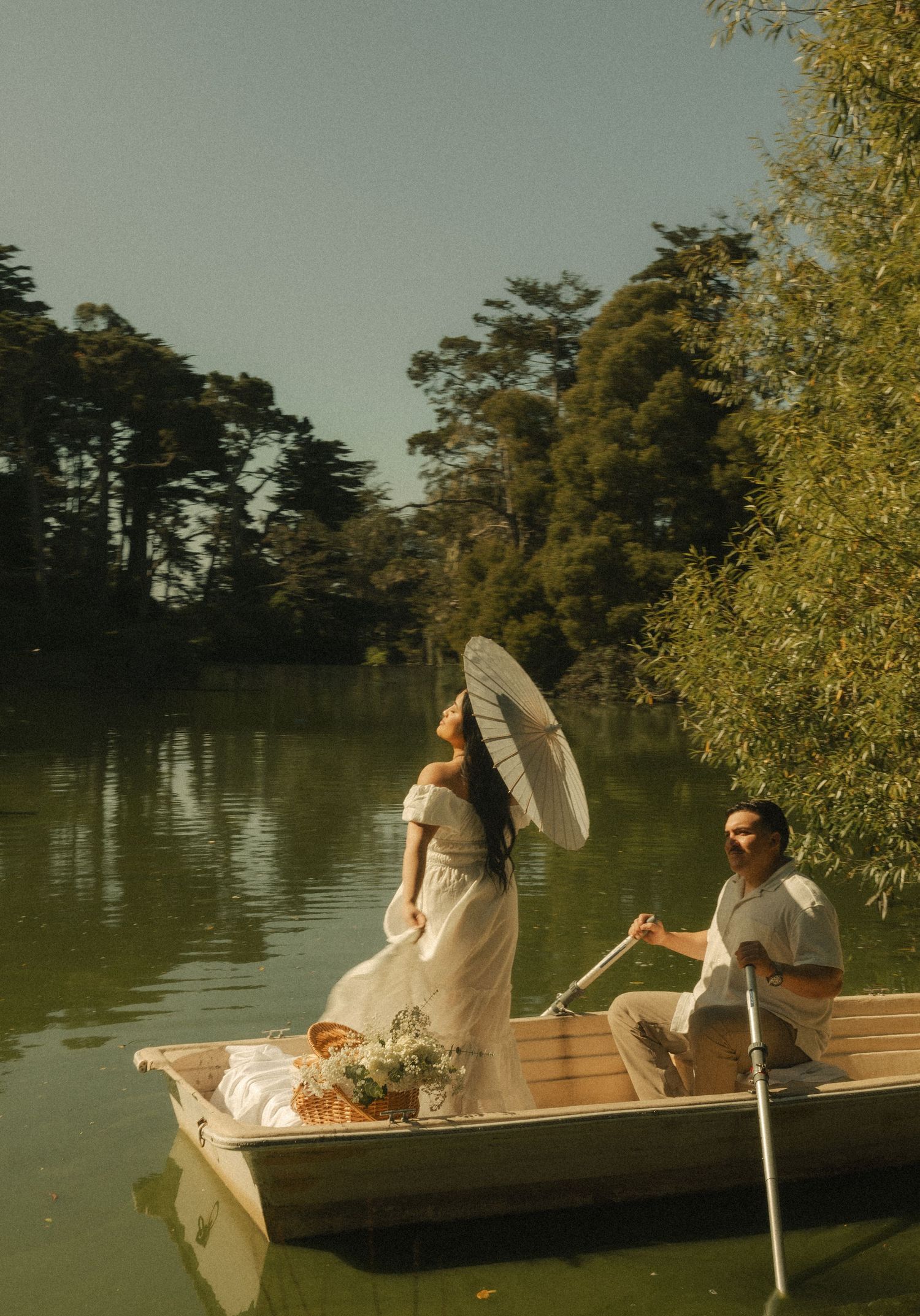 Couple on a wooden rowboat on a serene lake, woman in white dress holding parasol, man rowing near lush green trees.
