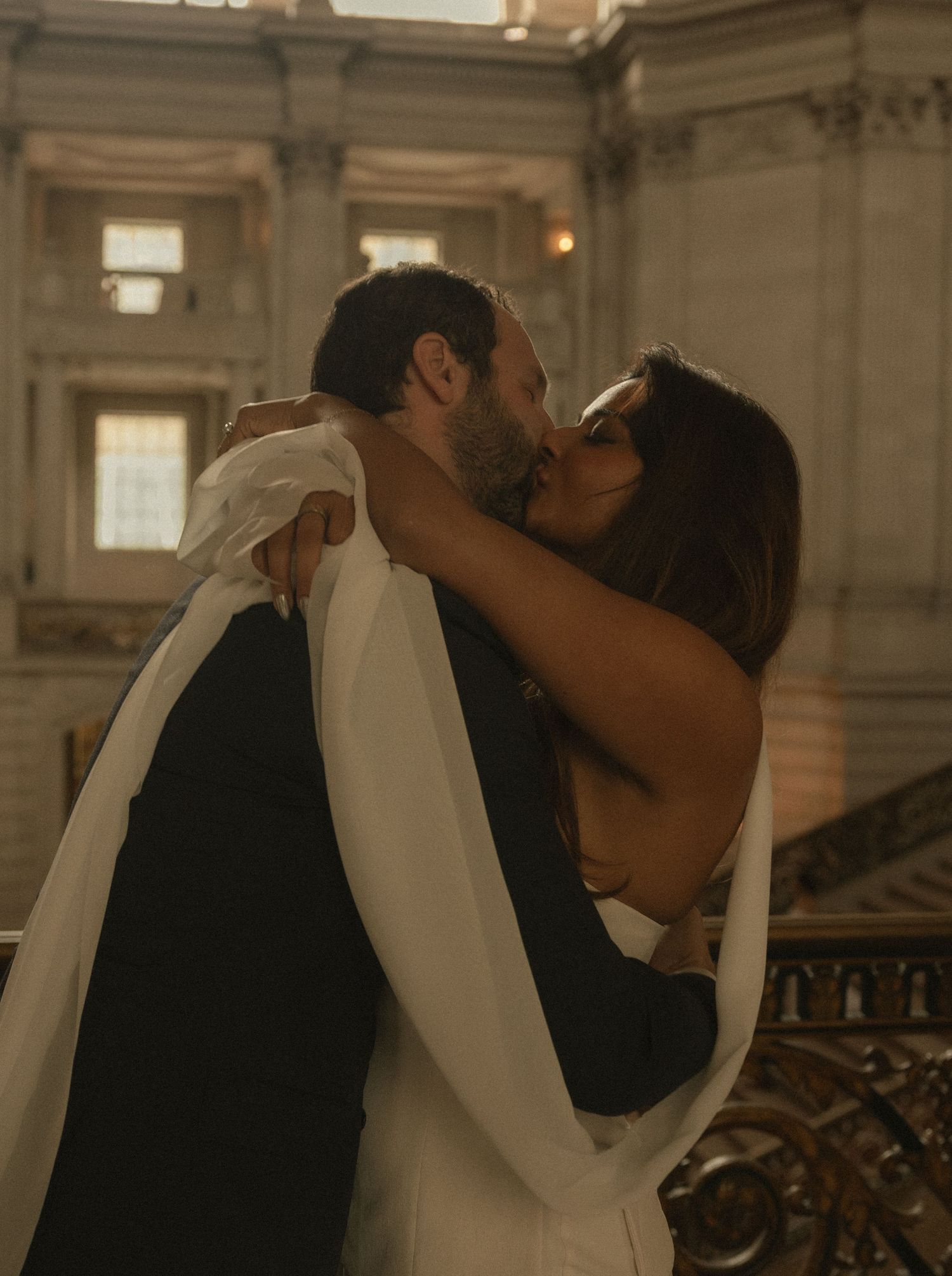 Couple sharing a romantic kiss at a wedding in an elegant grand hall with ornate architecture.