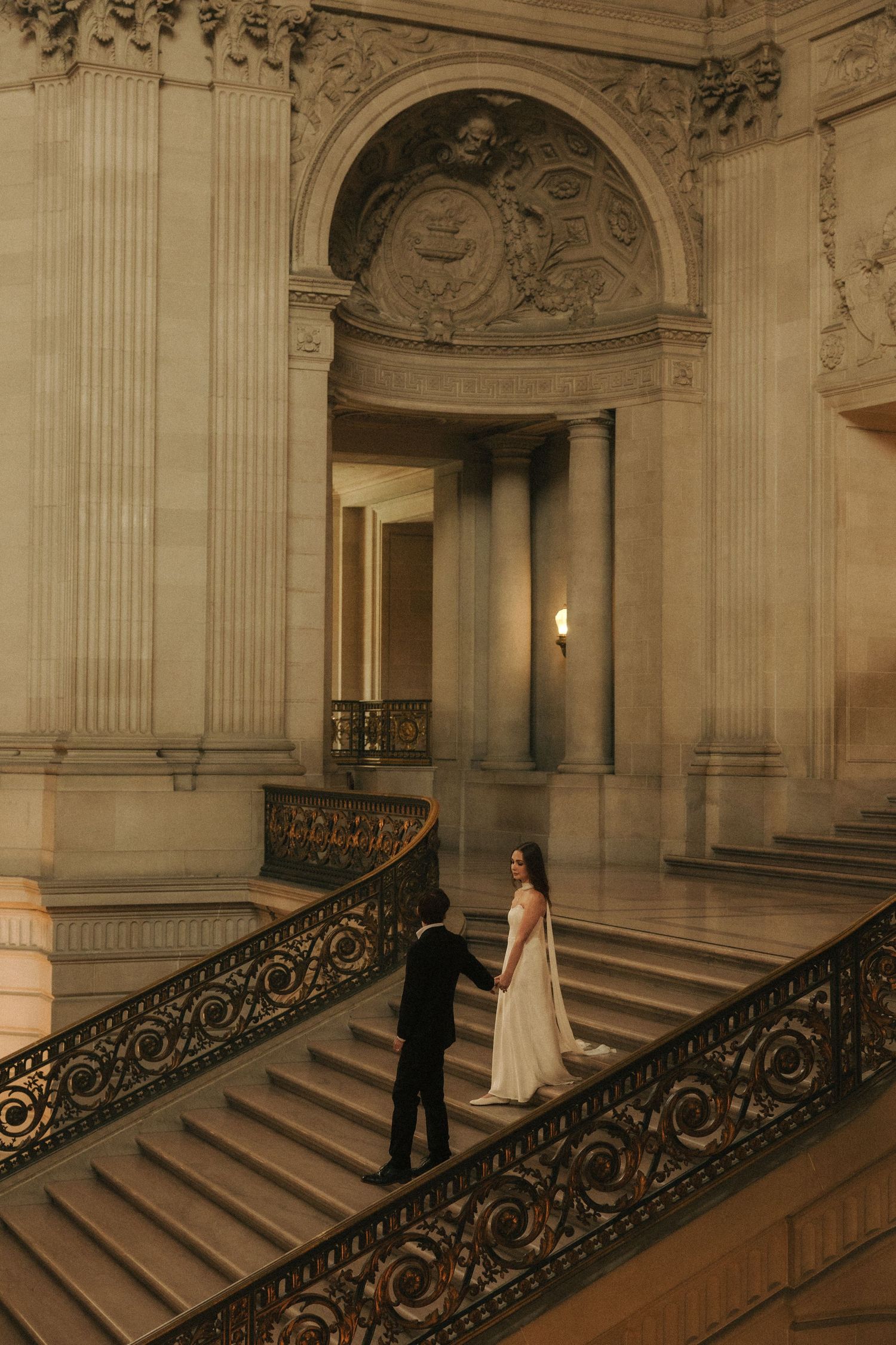 Couple in wedding attire holding hands on grand staircase inside ornate classical building with decorative ironwork.