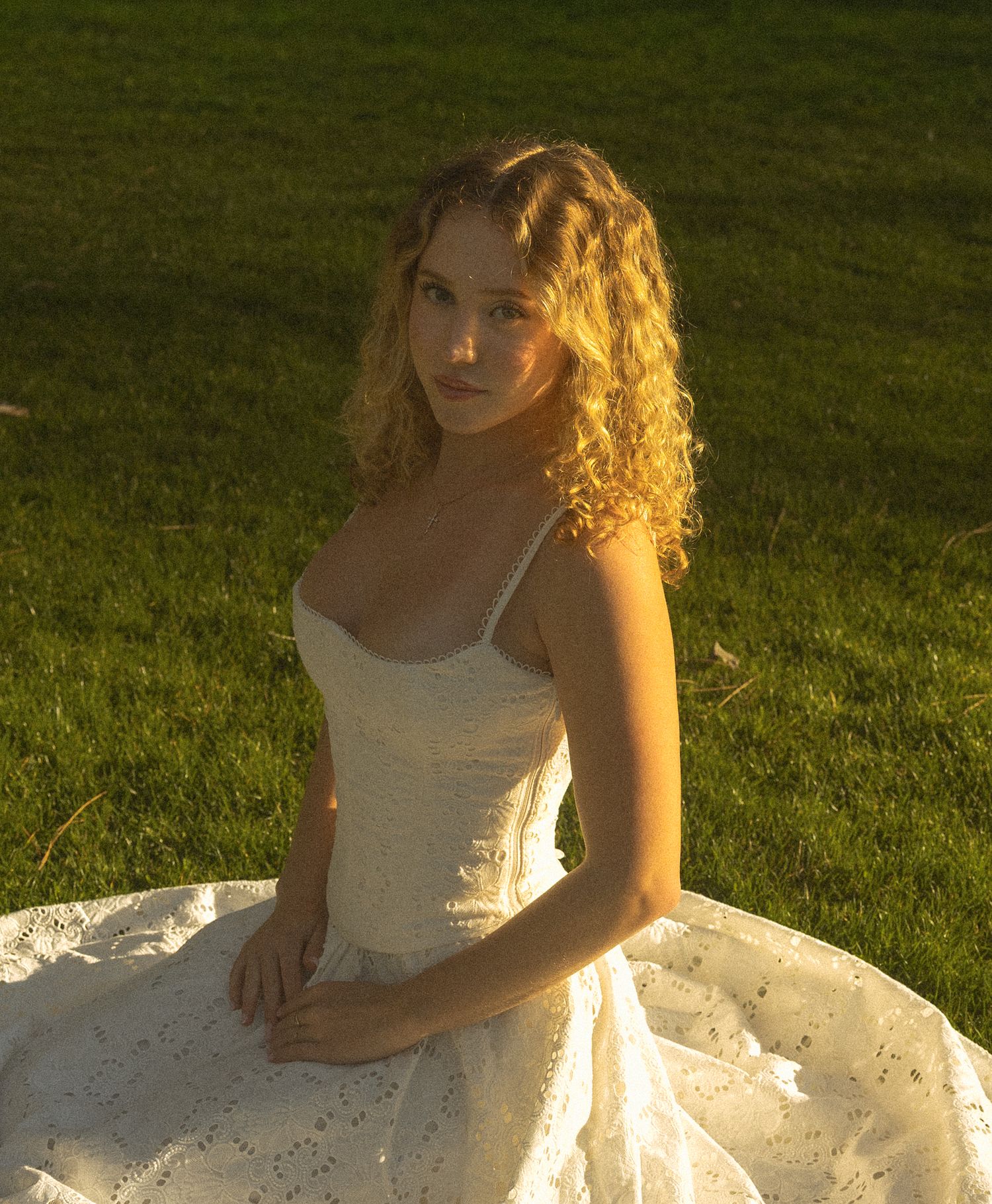 A figure in a white dress sits gracefully on a blanket in the grass during golden hour lighting.