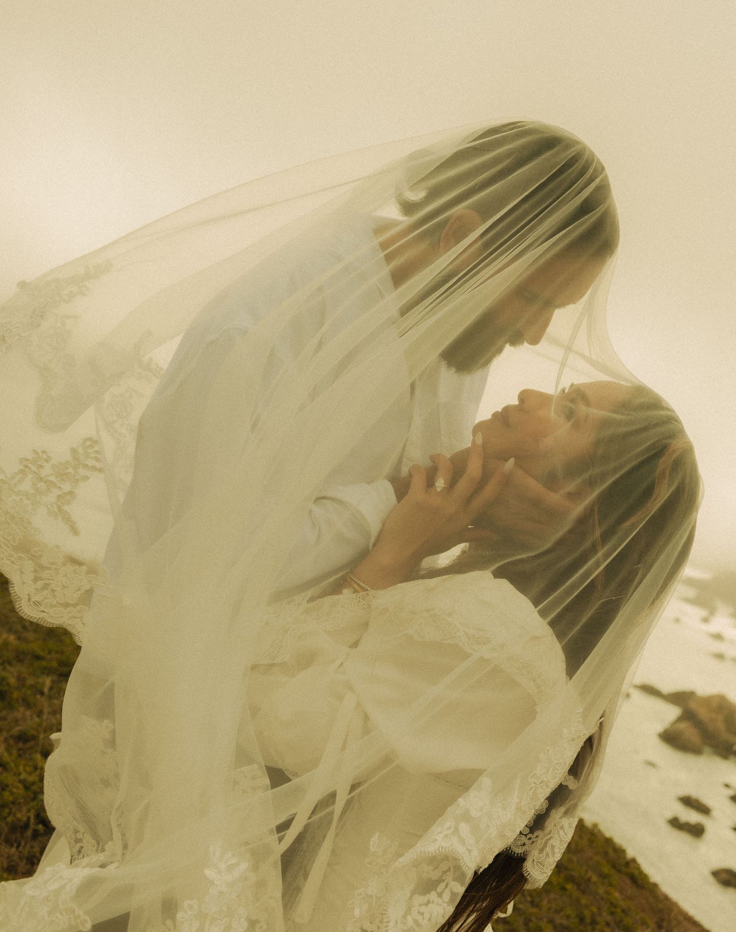 A bride and groom share an intimate moment under a flowing white lace veil outdoors in a misty landscape.