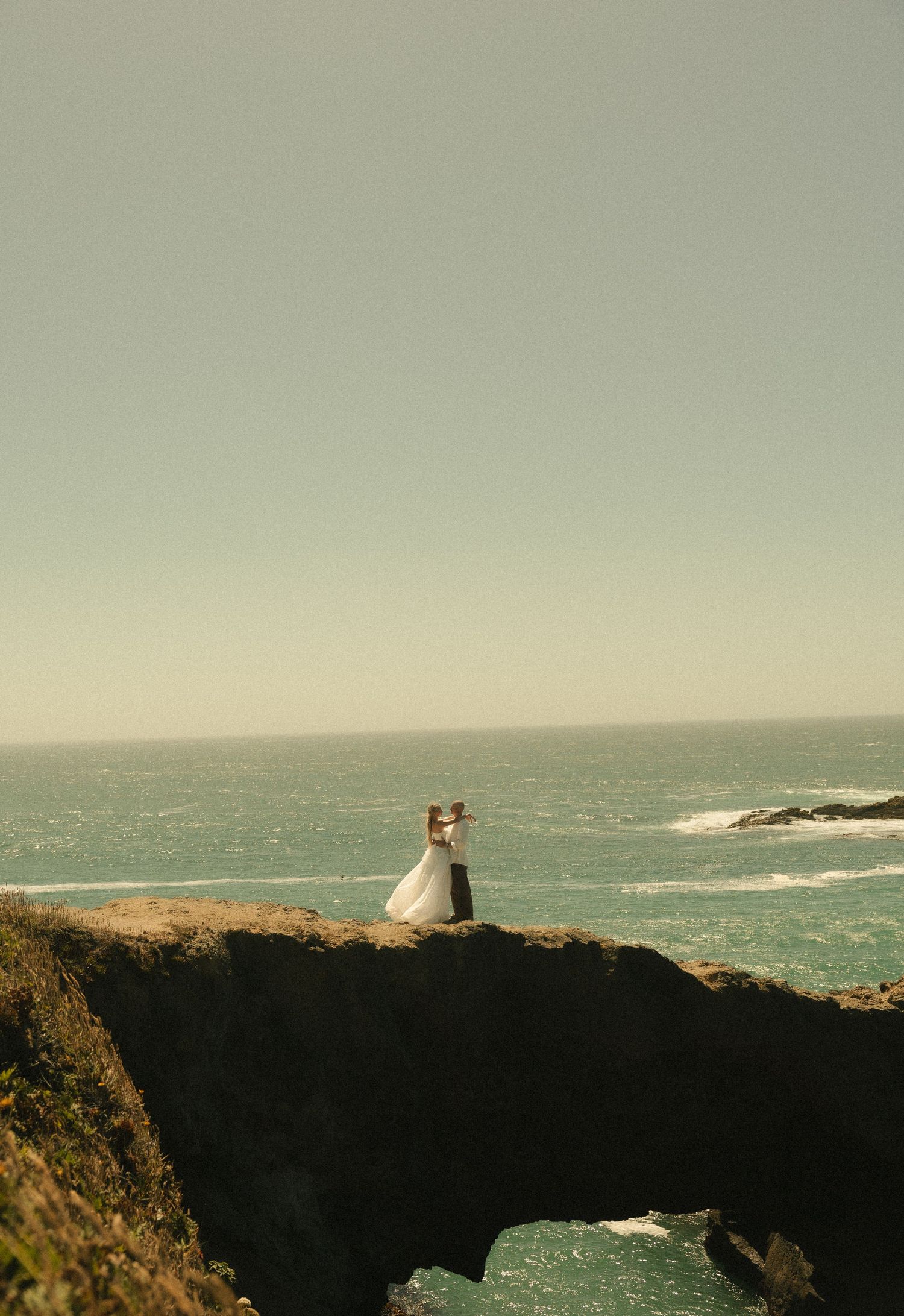 A bride and groom embrace on a rocky coastal cliff overlooking the ocean on their wedding day.