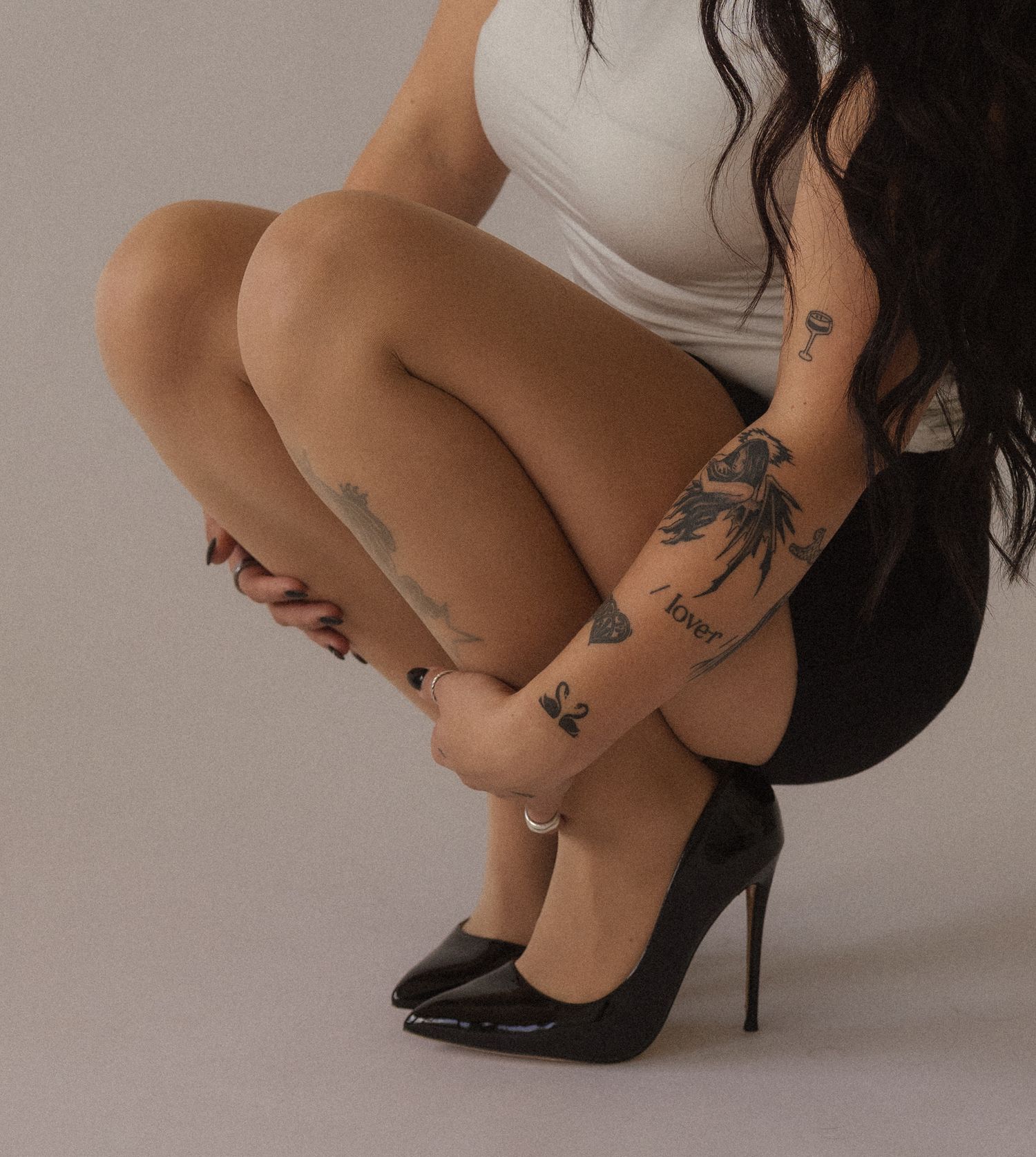 Woman squatting in black heels and white top, showcasing tattoos on her arms and legs against a gray background.