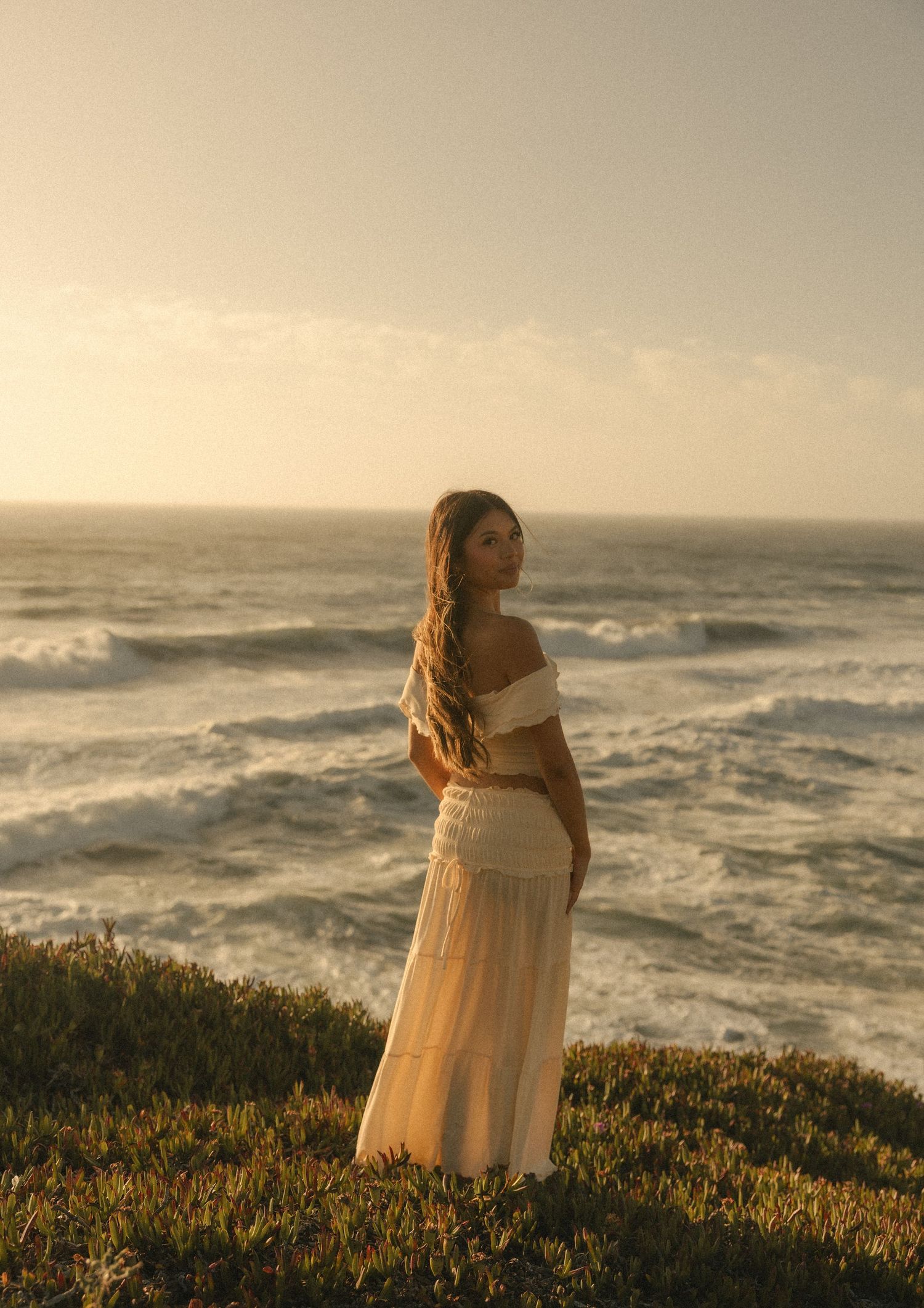 Woman in white off-shoulder maxi dress standing on coastal cliffside at golden hour with ocean waves behind her.