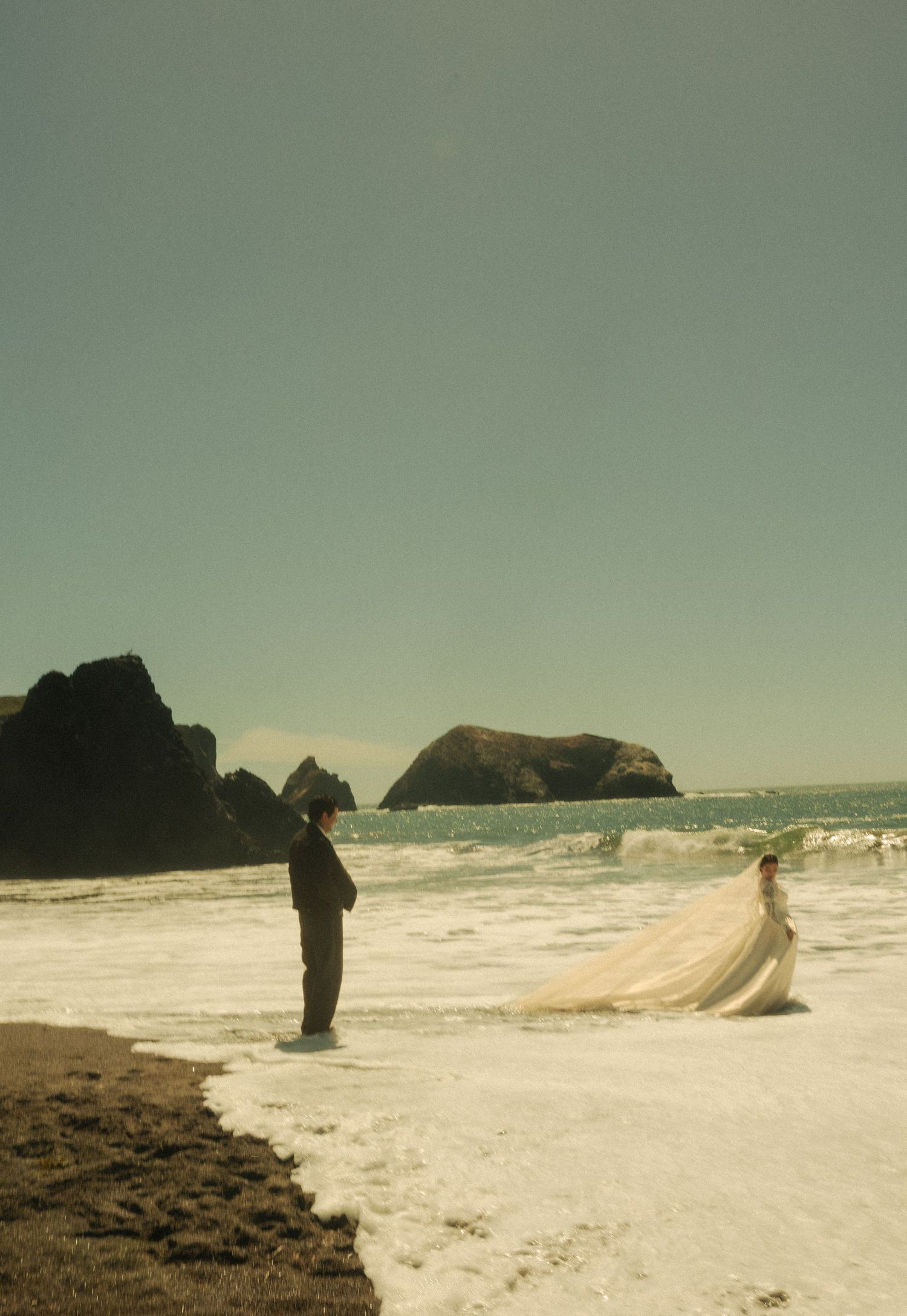 Bride in flowing white gown and groom stand on a misty beach with rocky sea stacks in the background.