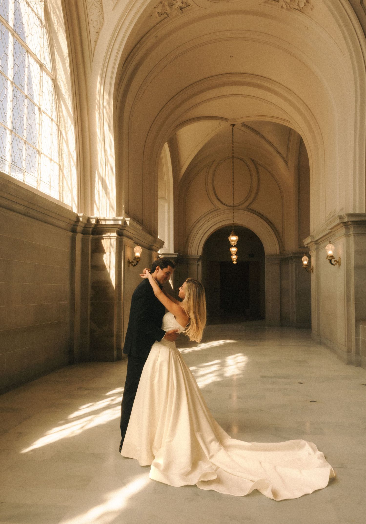 A bride and groom share a romantic dance in a grand arched hallway bathed in golden sunlight.