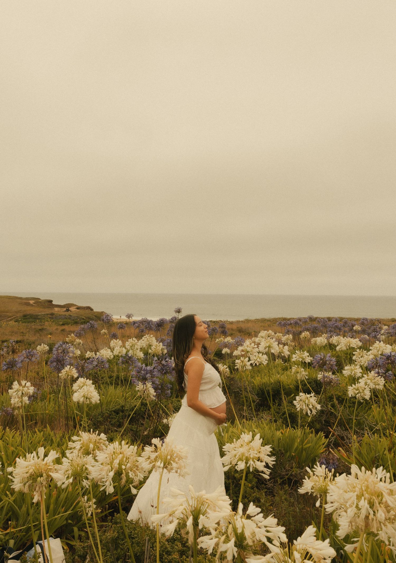 Pregnant woman in white dress standing among white and purple wildflowers on a coastal cliffside under an overcast sky.