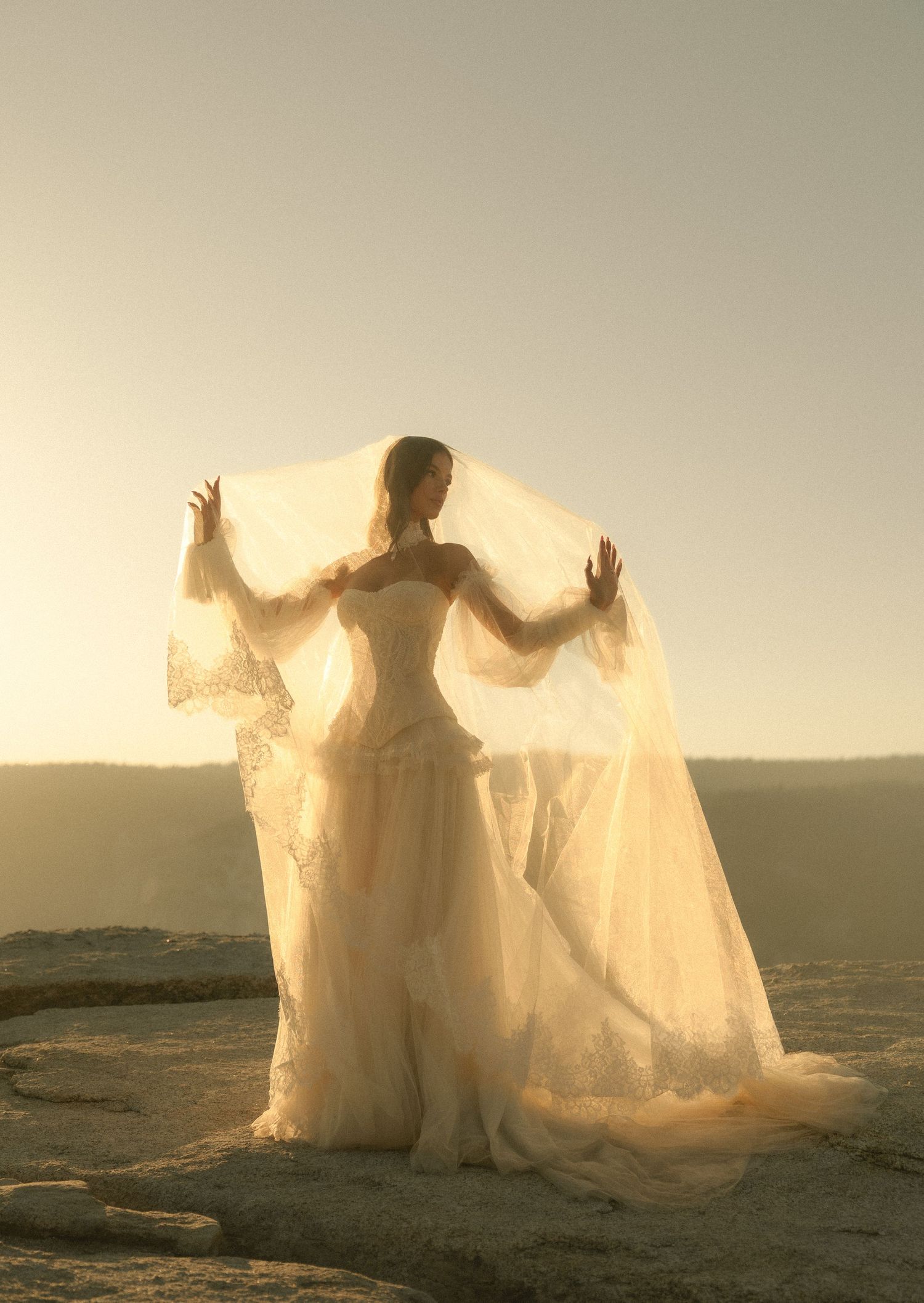 Bride in lace wedding gown holds flowing veil aloft on rocky cliff at golden sunset.