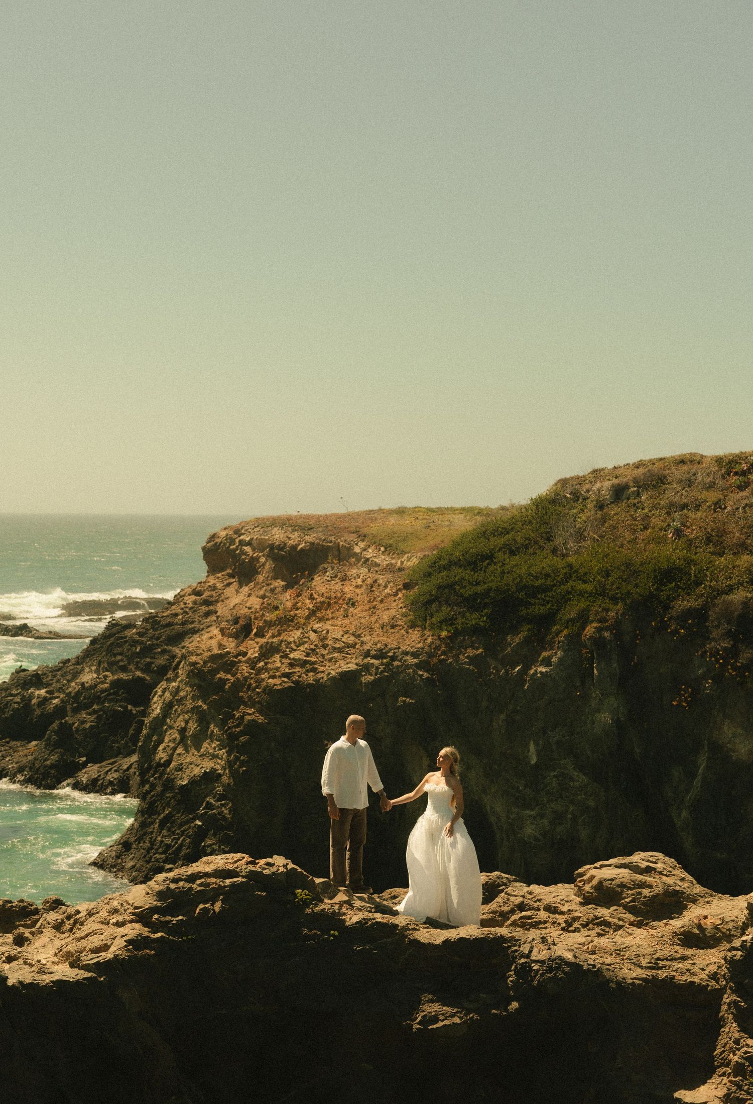 Bride and groom hold hands on rocky coastal cliffs overlooking the ocean during their romantic elopement ceremony.