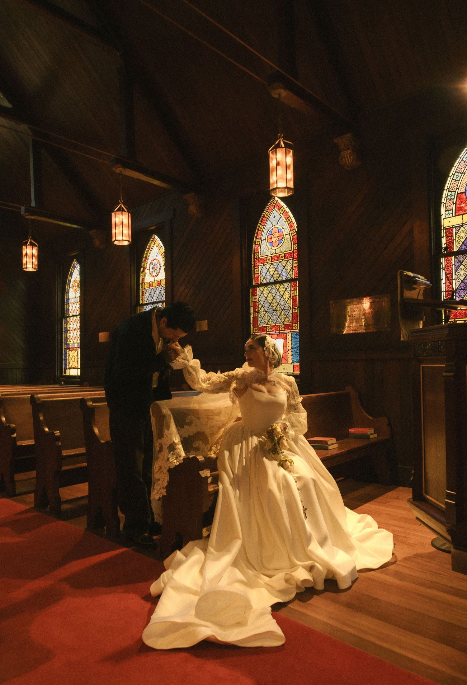 Bride and groom share a tender moment in a historic church with stunning stained glass windows and red carpet aisle.