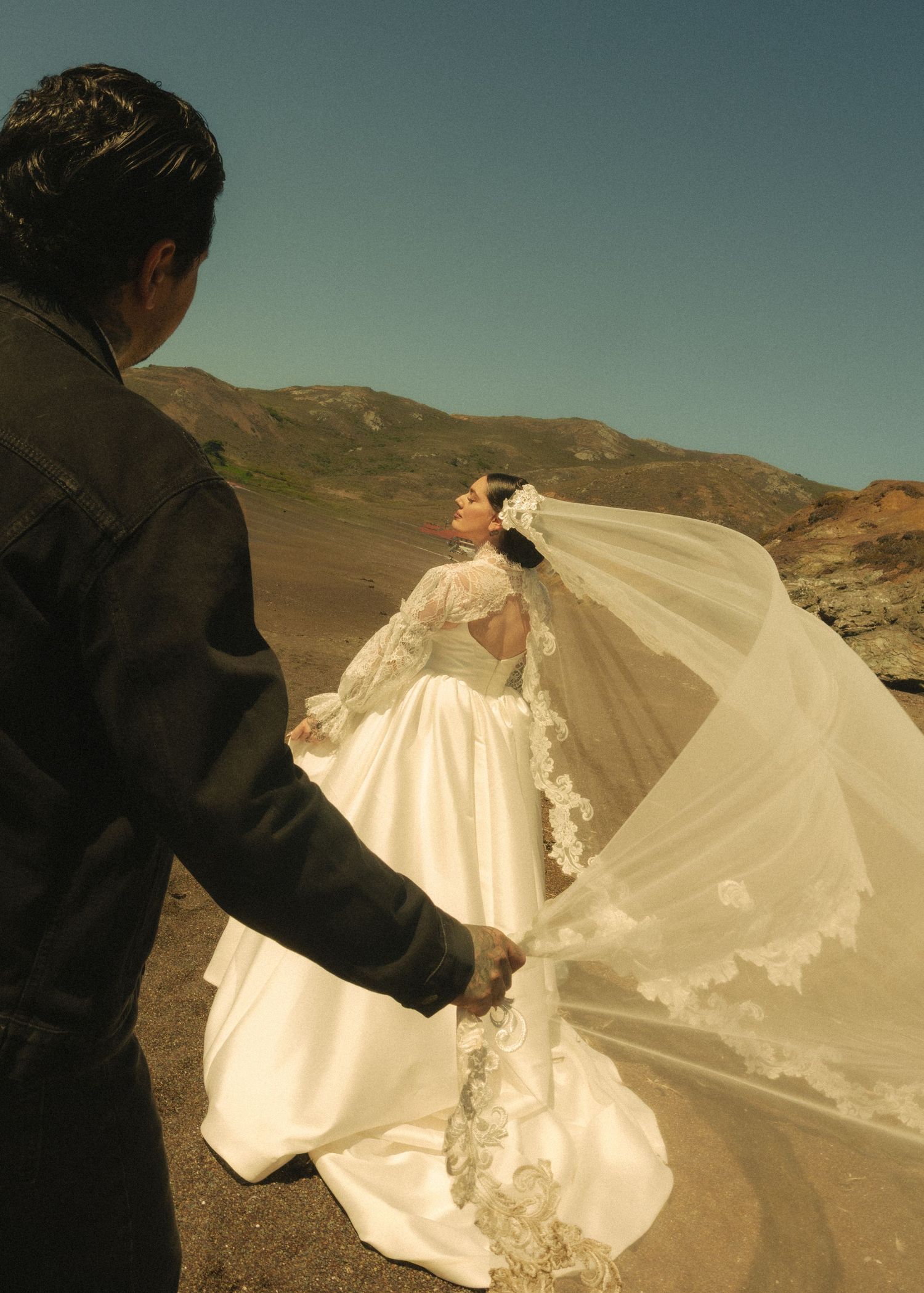 A bride in a flowing white gown and lace veil holds hands with a man in a black jacket against a desert mountain backdrop.