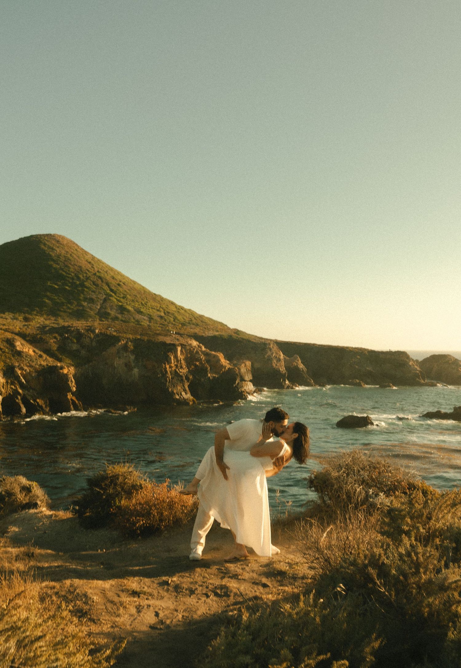 Couple sharing a romantic kiss on a coastal cliff at sunset with ocean waves and green hills in Big Sur, California.