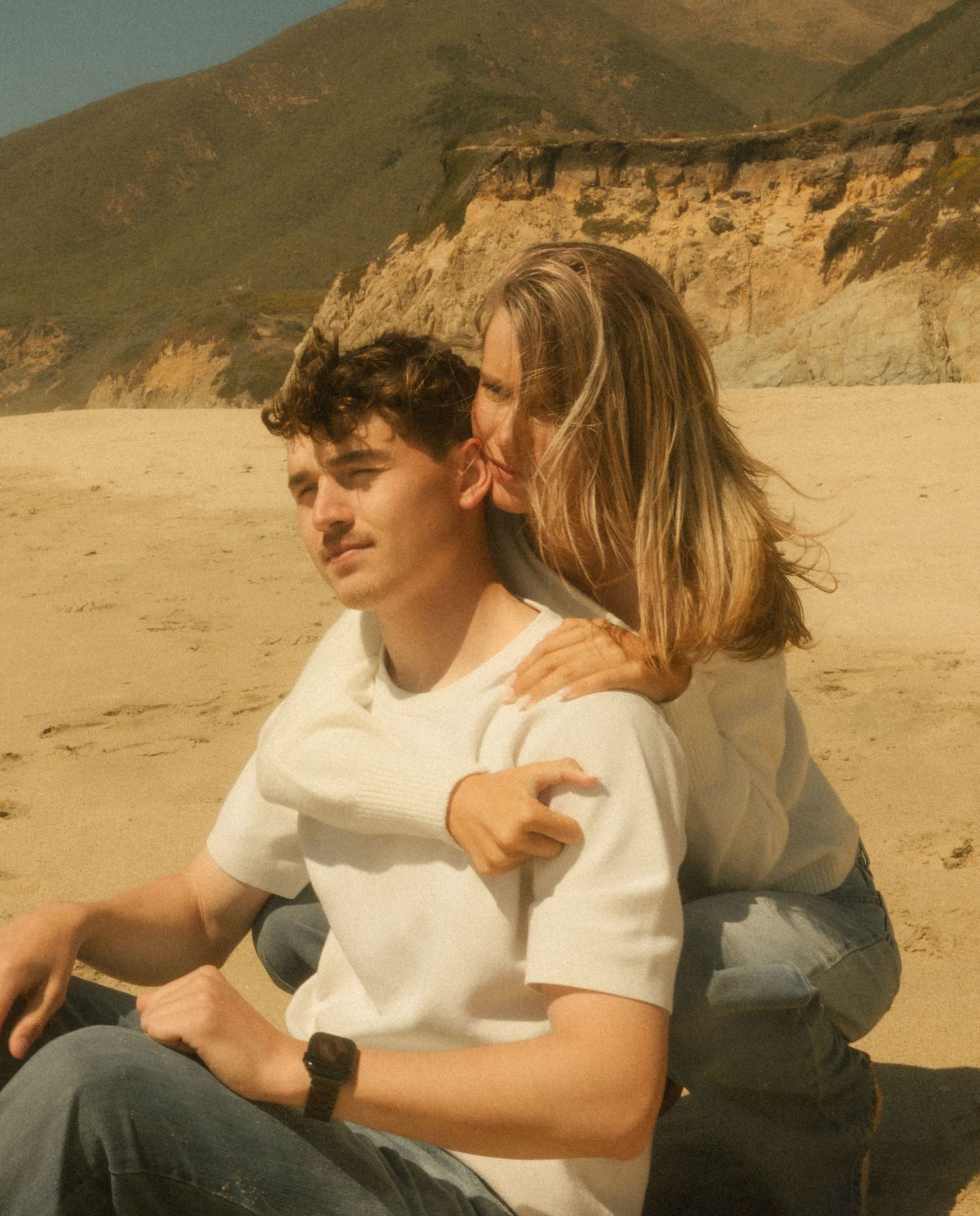 A young couple sits together on a sandy beach with rocky cliffs, the woman embracing the man from behind.