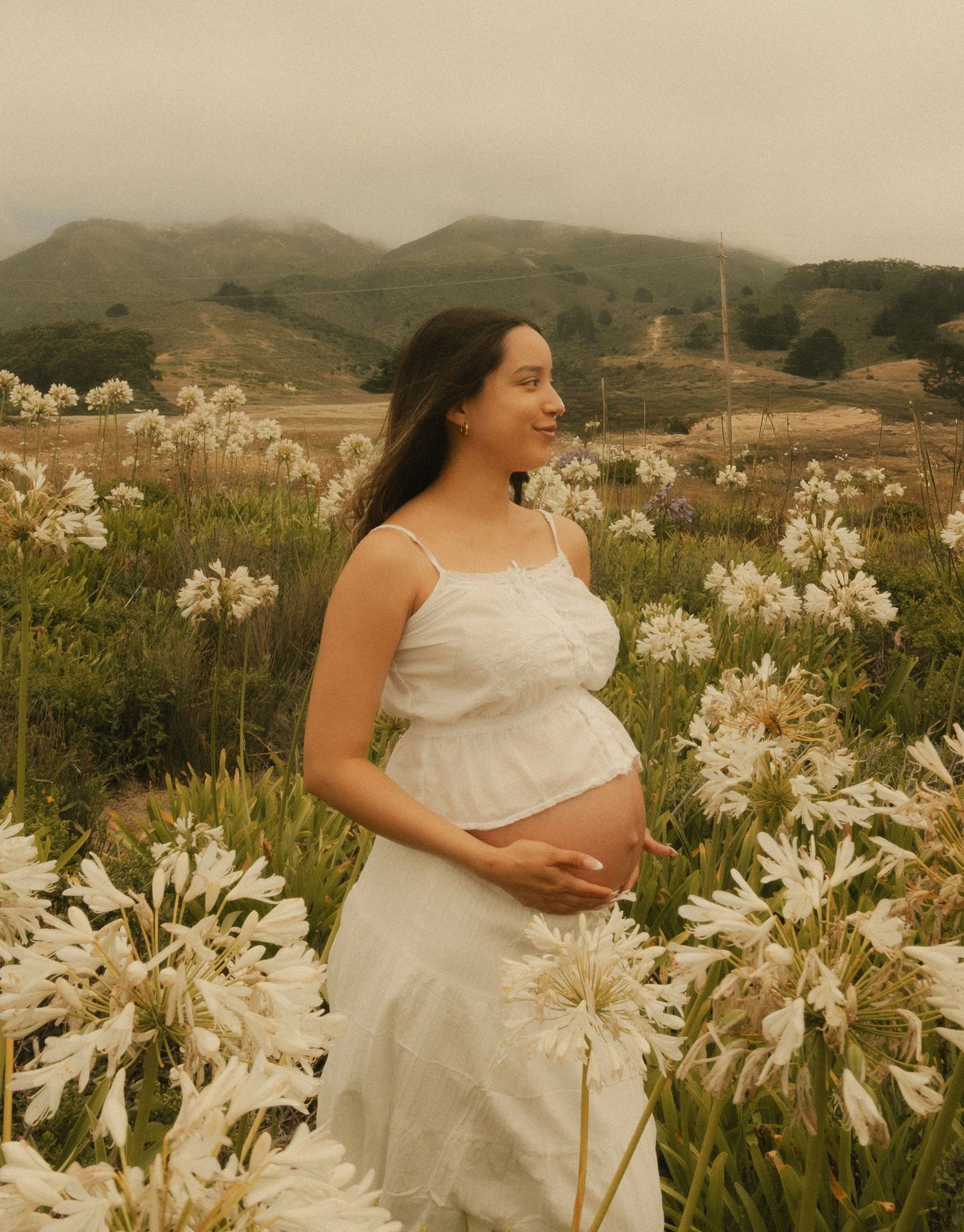 Pregnant woman in white dress cradling baby bump in a field of white agapanthus flowers with misty mountains behind.