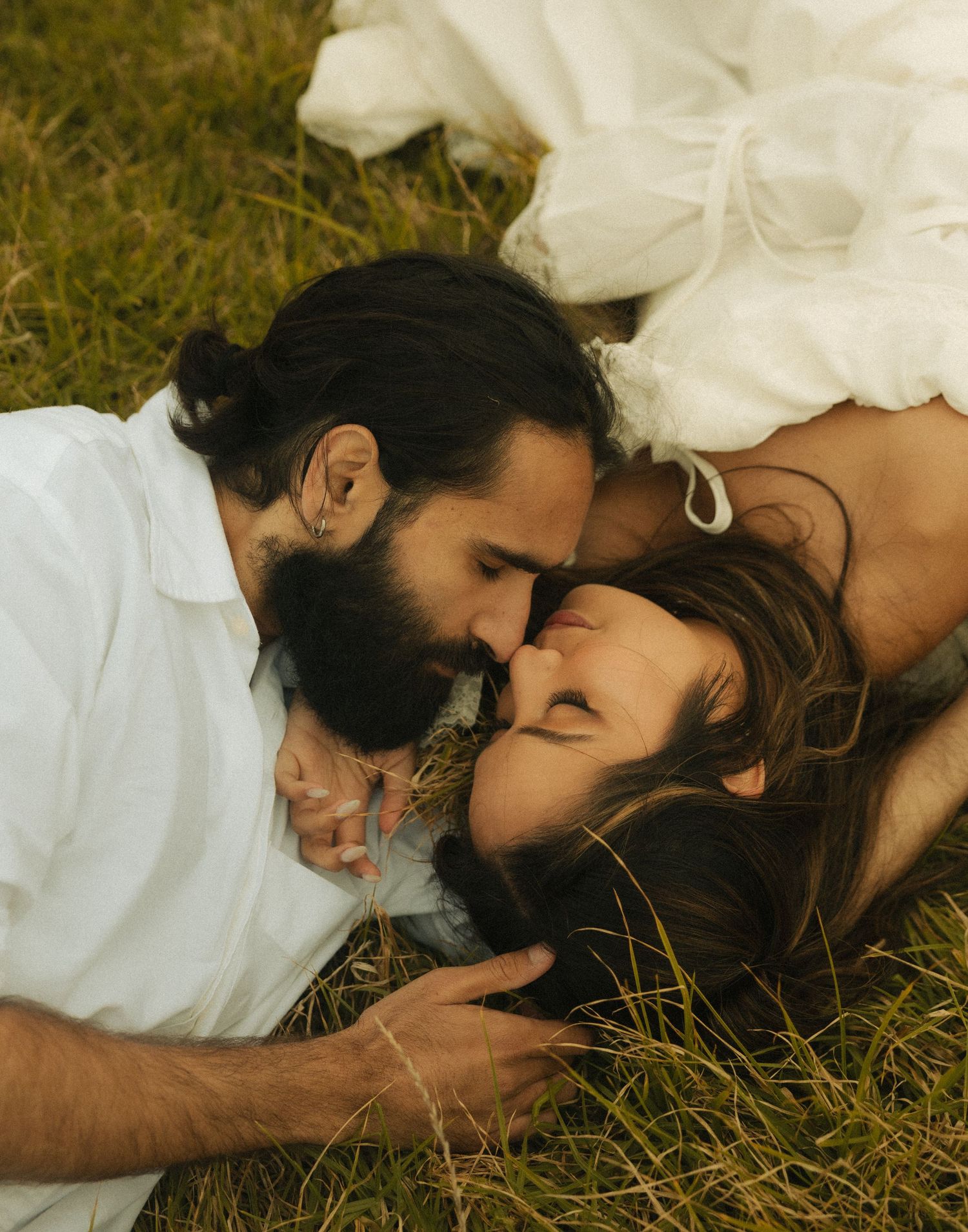 A bearded groom and bride in white dress lying in grass, sharing a tender romantic kiss on their wedding day.
