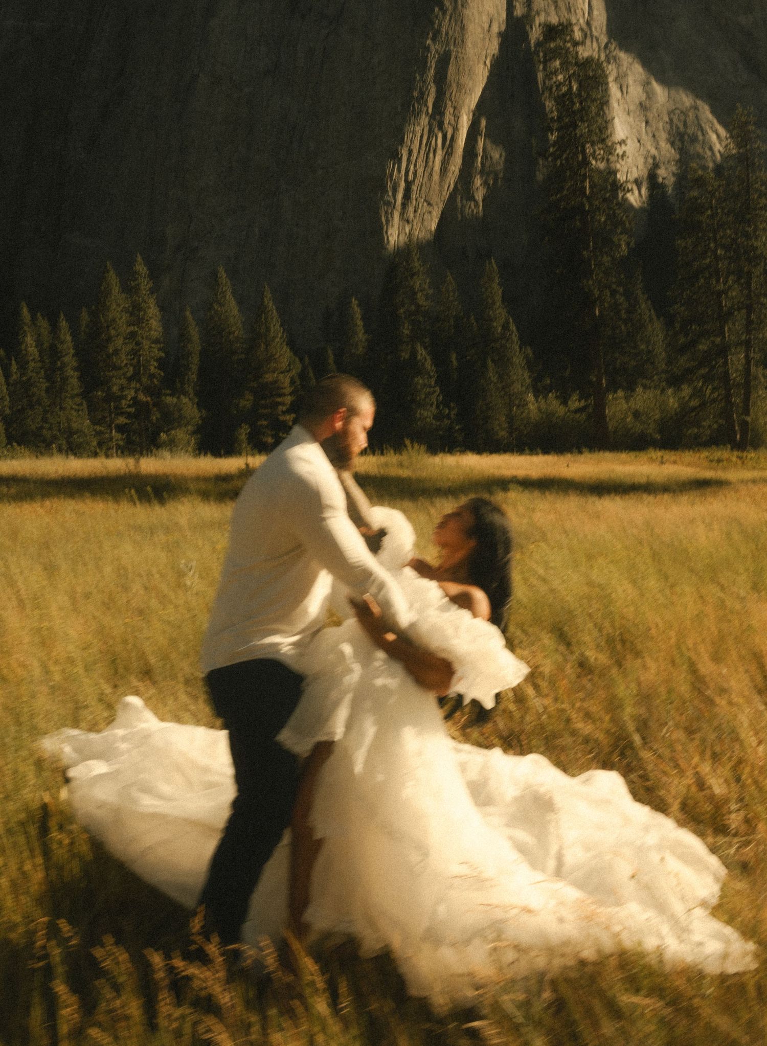 Couple dancing in a golden meadow with dramatic cliffs and pine trees in Yosemite, bride in flowing white gown.