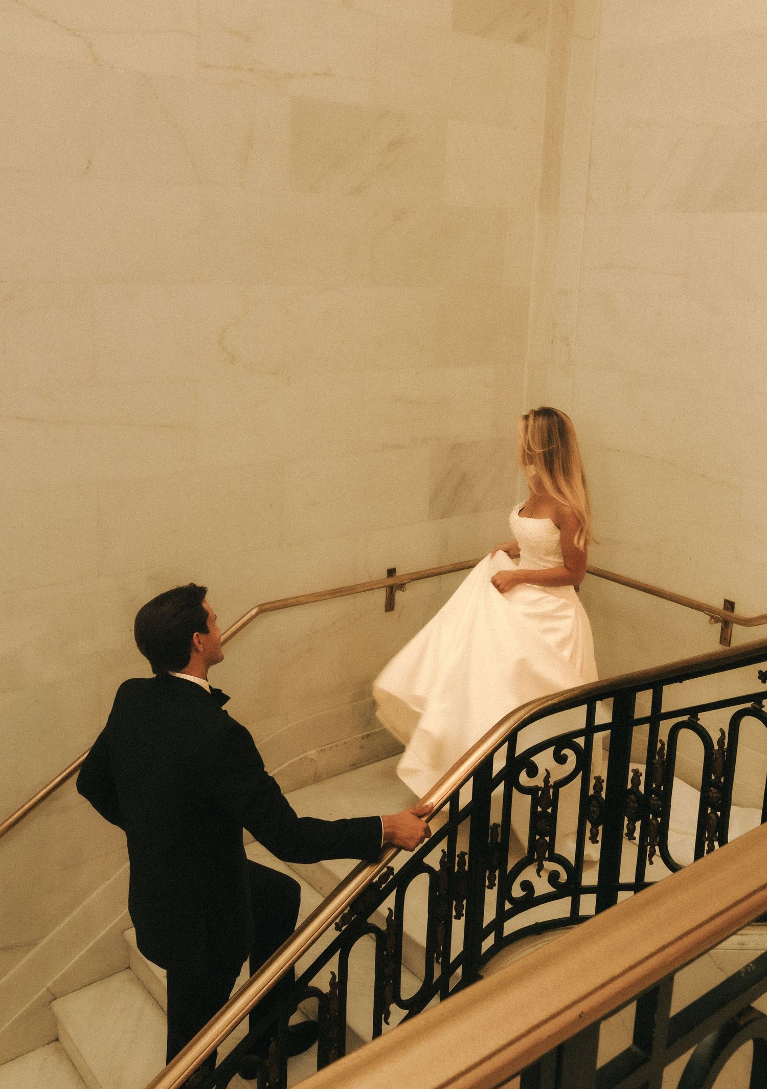 Bride and groom hold hands on elegant marble staircase with wrought iron railing, bride in white gown looks back.