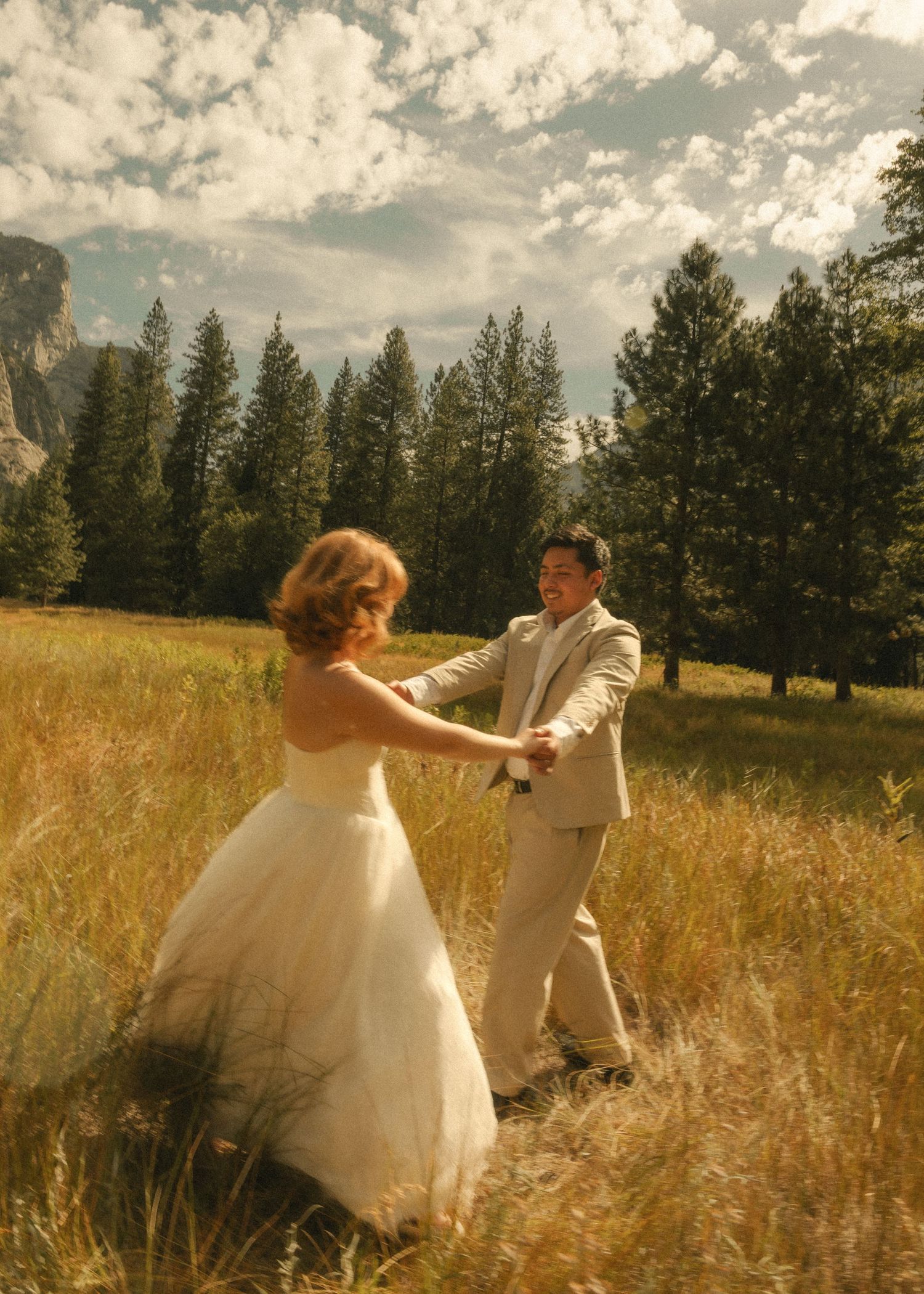 Bride and groom dancing in golden meadow with pine trees and mountains under cloudy sky at Yosemite wedding.