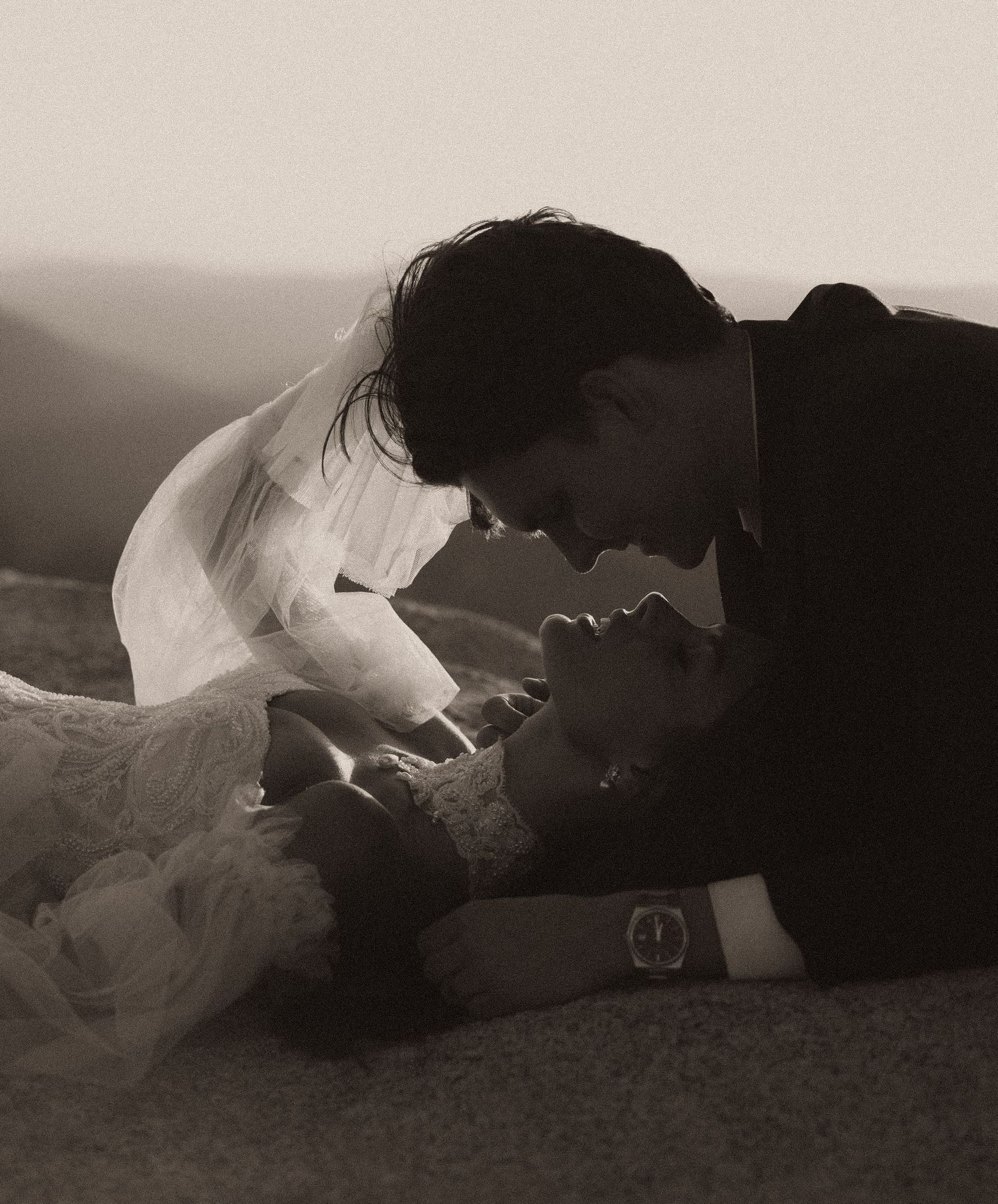 Black and white photo of a romantic couple sharing an intimate moment, bride in lace veil lying down with groom leaning close.
