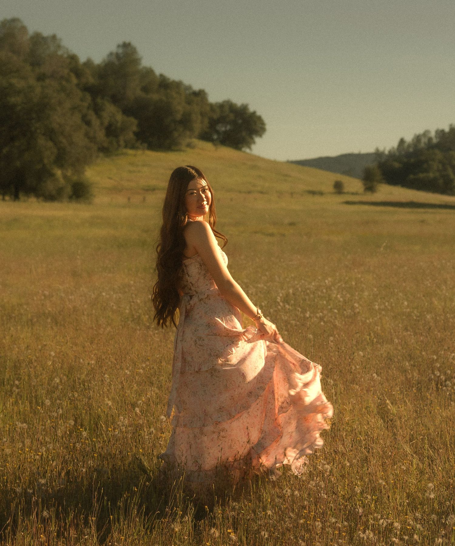 Woman in flowing pink floral dress twirling in golden meadow at sunset with green hills behind her.