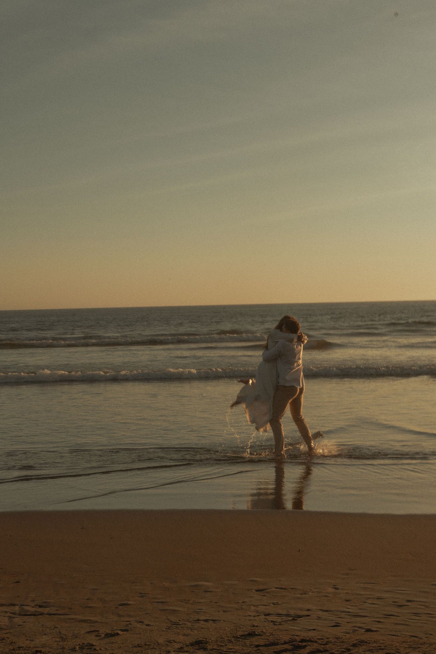 Two figures in white dresses dance playfully along a sunset beach with ocean waves in the background.