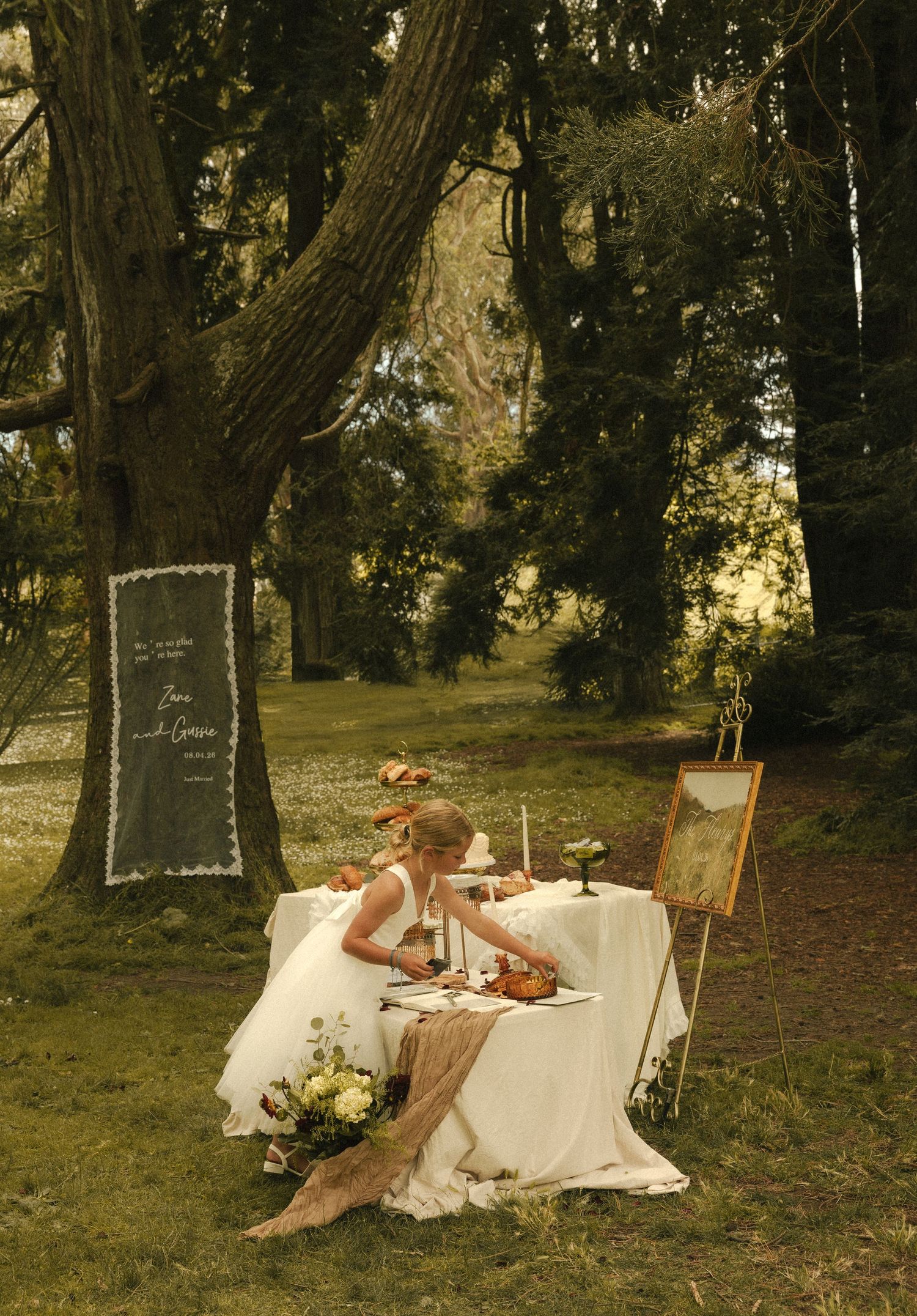 Bride in white gown seated at elegant outdoor sweetheart table surrounded by lush trees and floral decor.