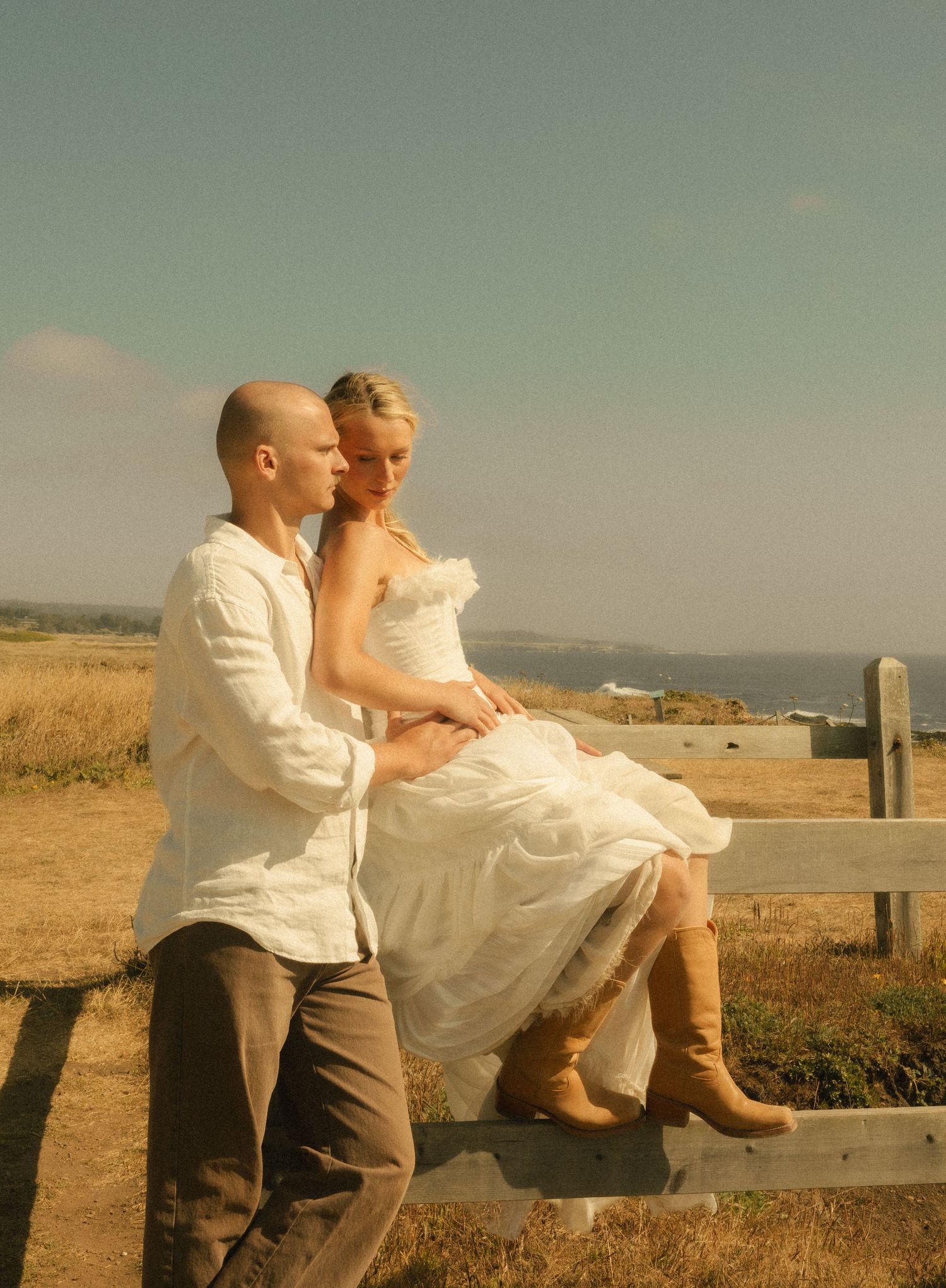 Bride in white dress and cowboy boots sits on fence with groom in white shirt, ocean backdrop on sunny day.