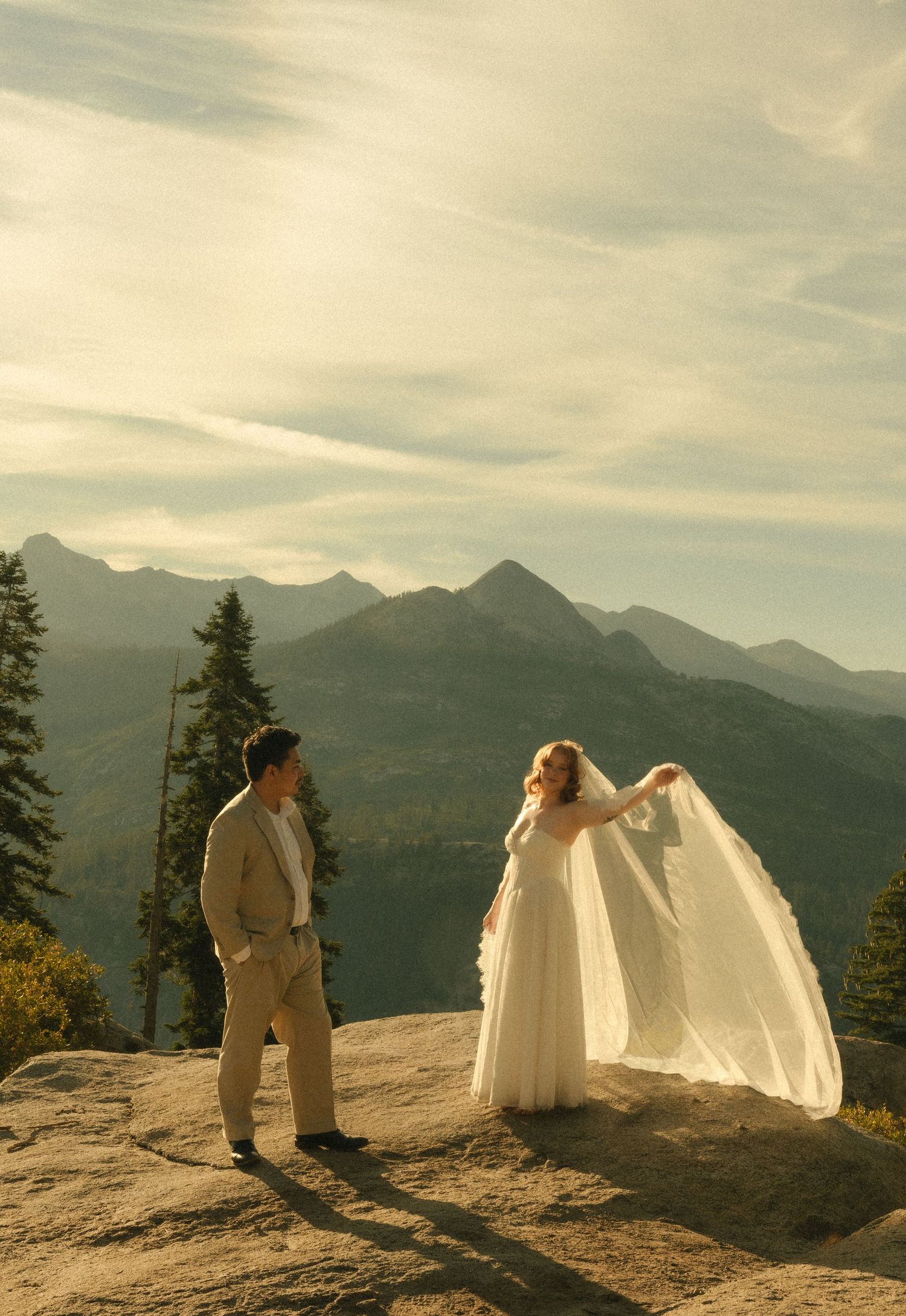 Bride in flowing white gown and groom in tan suit pose on mountain summit with scenic peaks backdrop.
