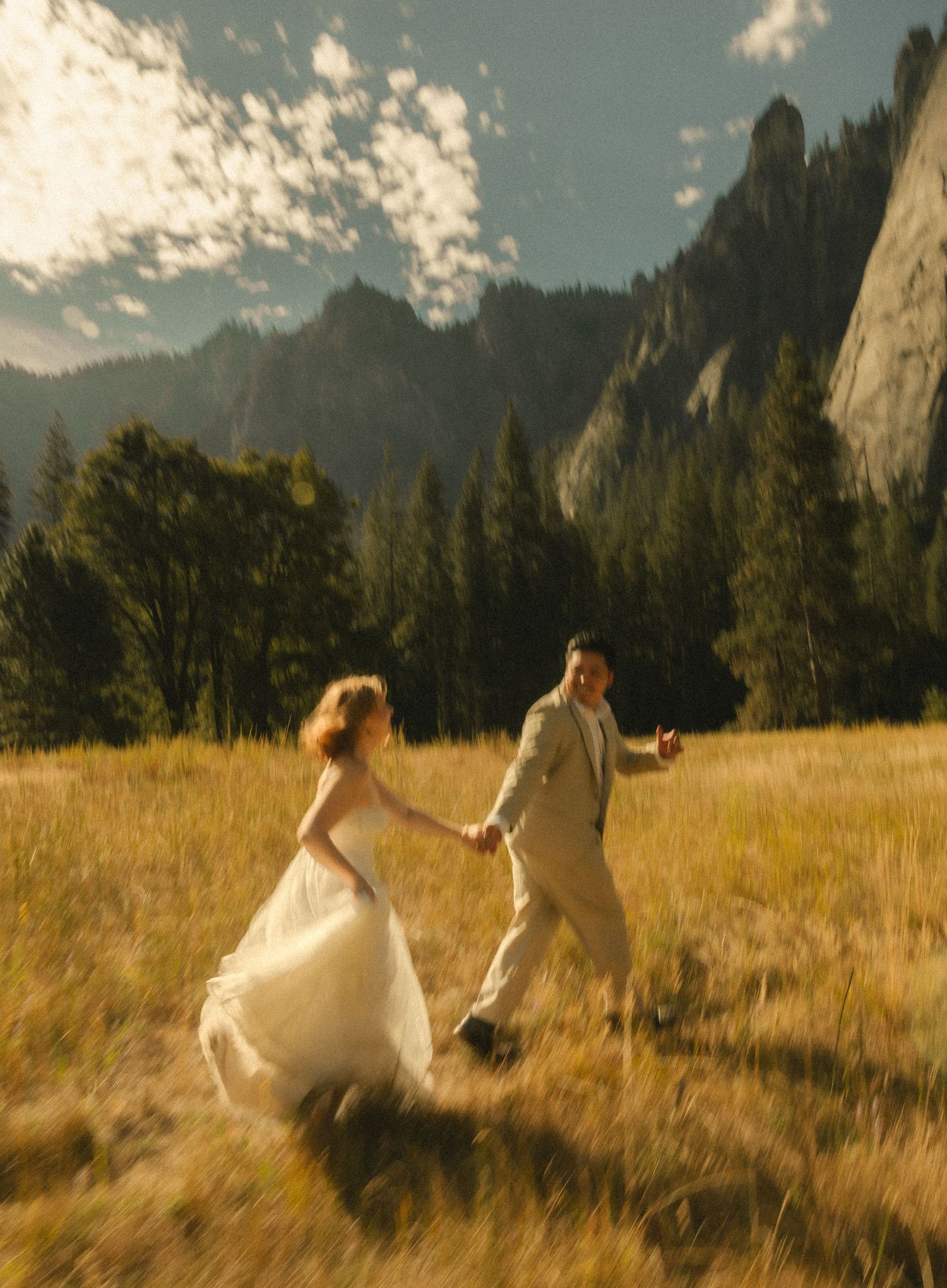 A bride and groom run hand-in-hand through a golden meadow with dramatic mountain cliffs and pine trees behind them.