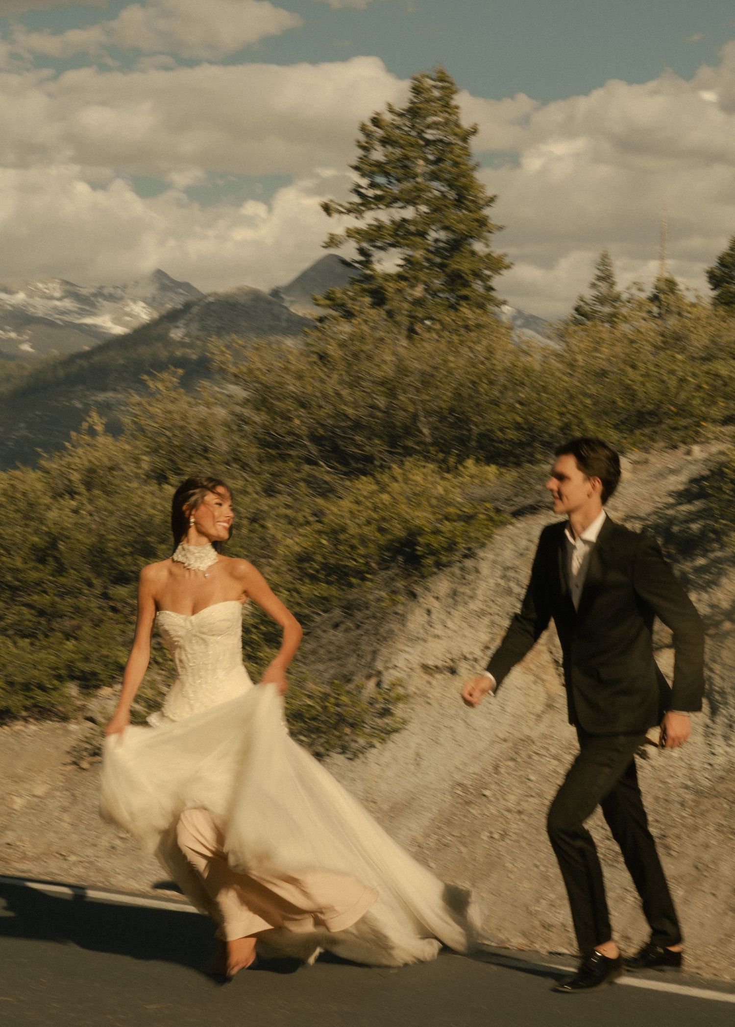 Bride in flowing white gown and groom in black suit walk hand-in-hand on mountain road with scenic peaks backdrop.