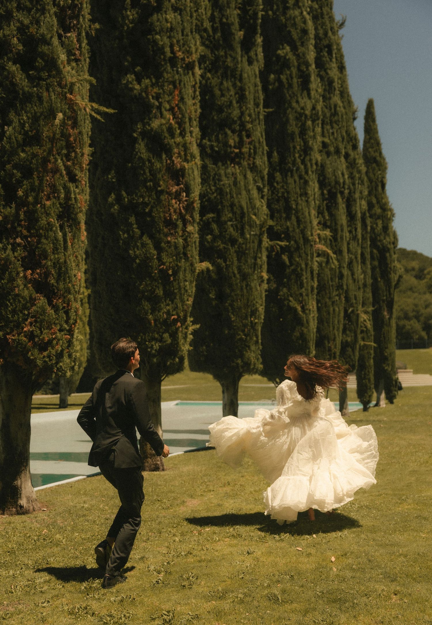 A bride in a flowing white gown and groom in black suit run playfully on green lawn beside tall cypress trees.