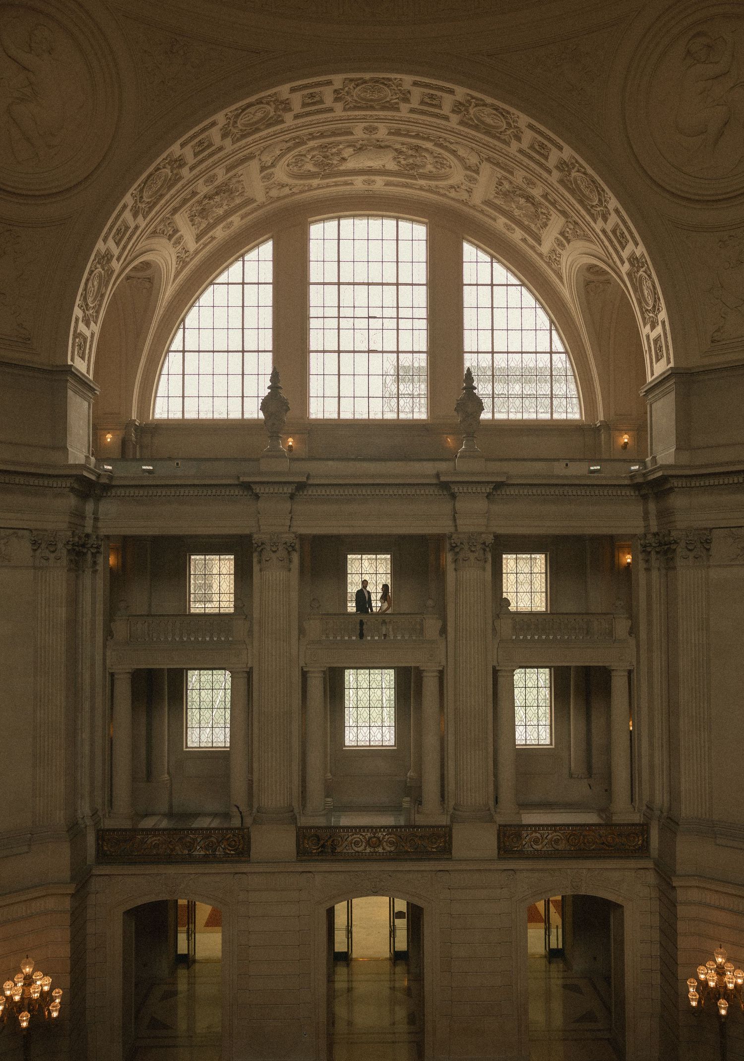 Grand neoclassical rotunda interior with arched window, ornate columns, and two figures standing on upper balcony.