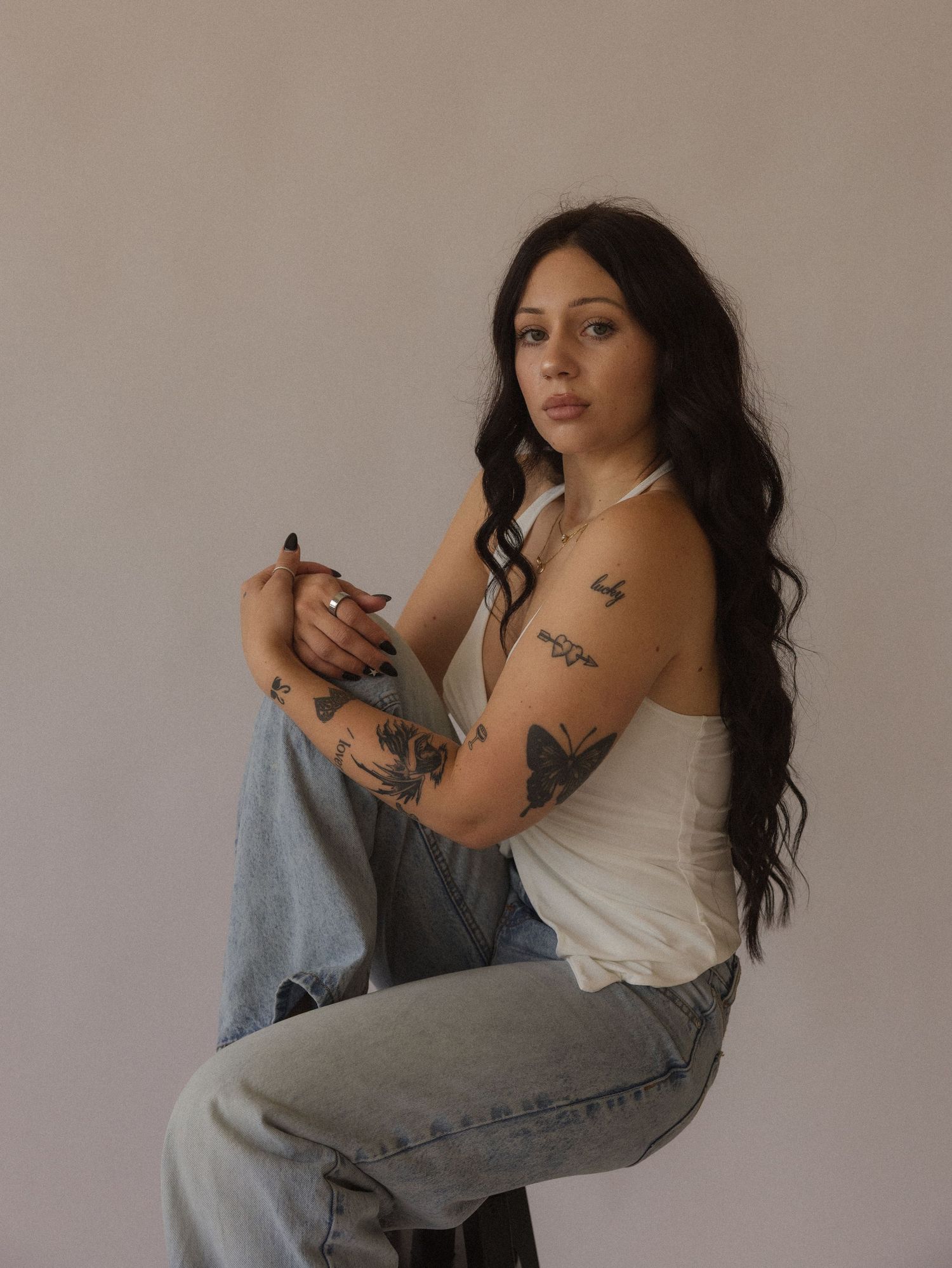 Young woman with long dark hair and tattoos sits on a stool wearing a white tank top and jeans.